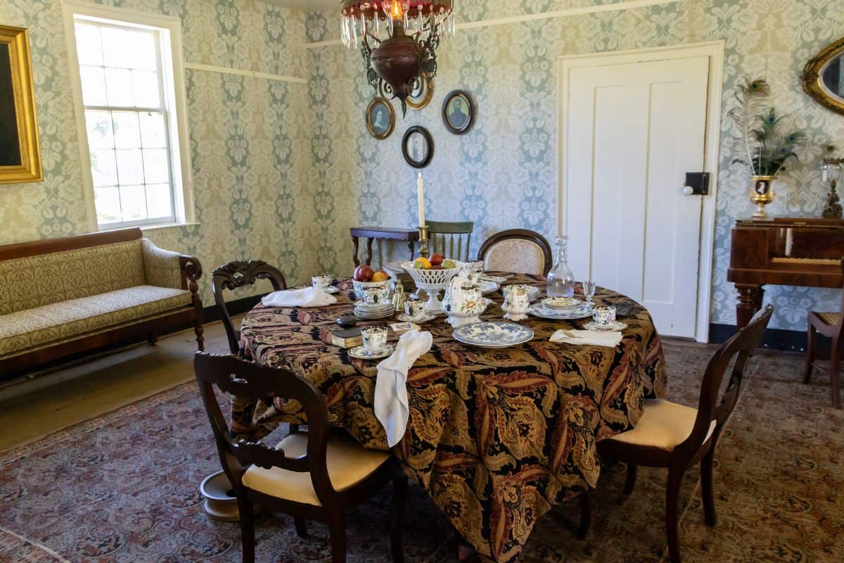 A dining room with a round table set for tea, patterned tablecloth, four chairs, a sofa, piano, and framed pictures on wallpapered walls.