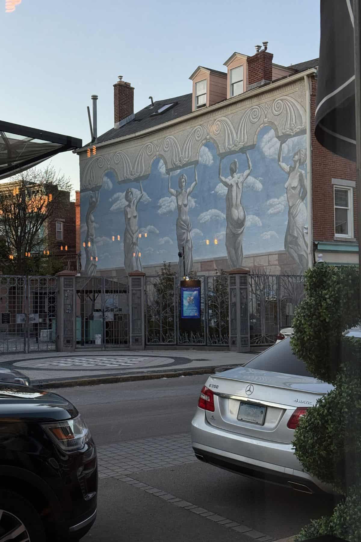 A mural of five women holding a patterned cloth above their heads is painted on the side of a building above a gated courtyard; cars are parked in front.