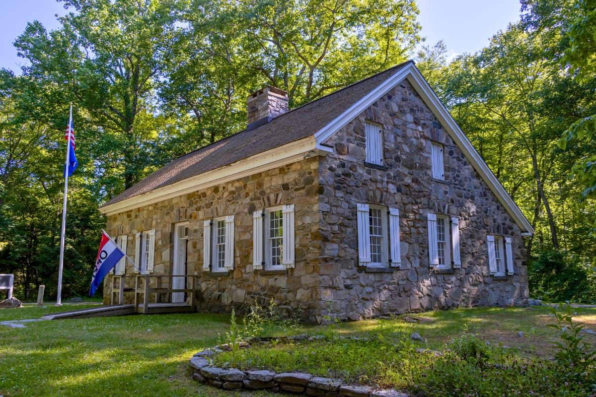 A historic stone house with white-trimmed windows stands among trees. Two flags, including an "OPEN" flag, are displayed near the entrance.