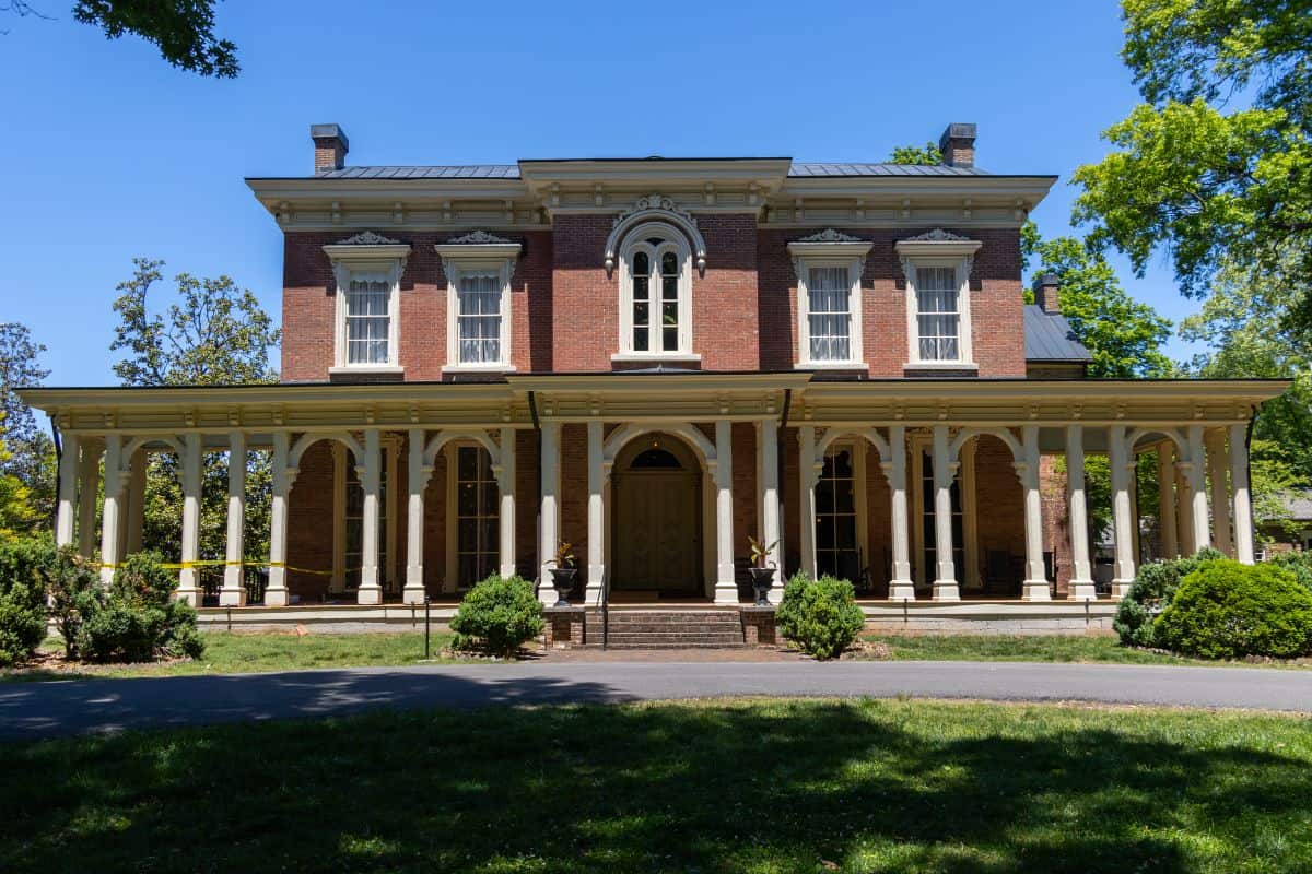 A two-story brick house with tall windows, white trim, and a large covered porch supported by columns, surrounded by trees and shrubs on a sunny day.