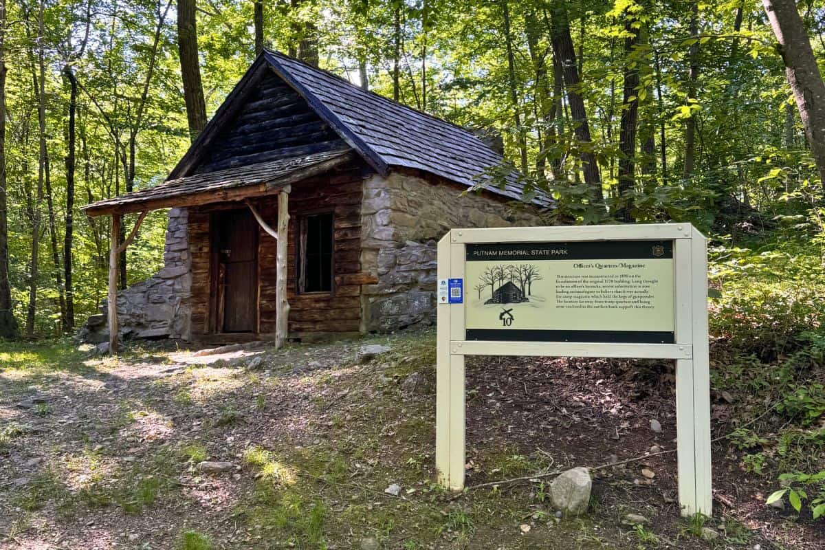 A small stone cabin with a wooden roof stands in a forest next to a large informational sign for Putnam Memorial State Park.