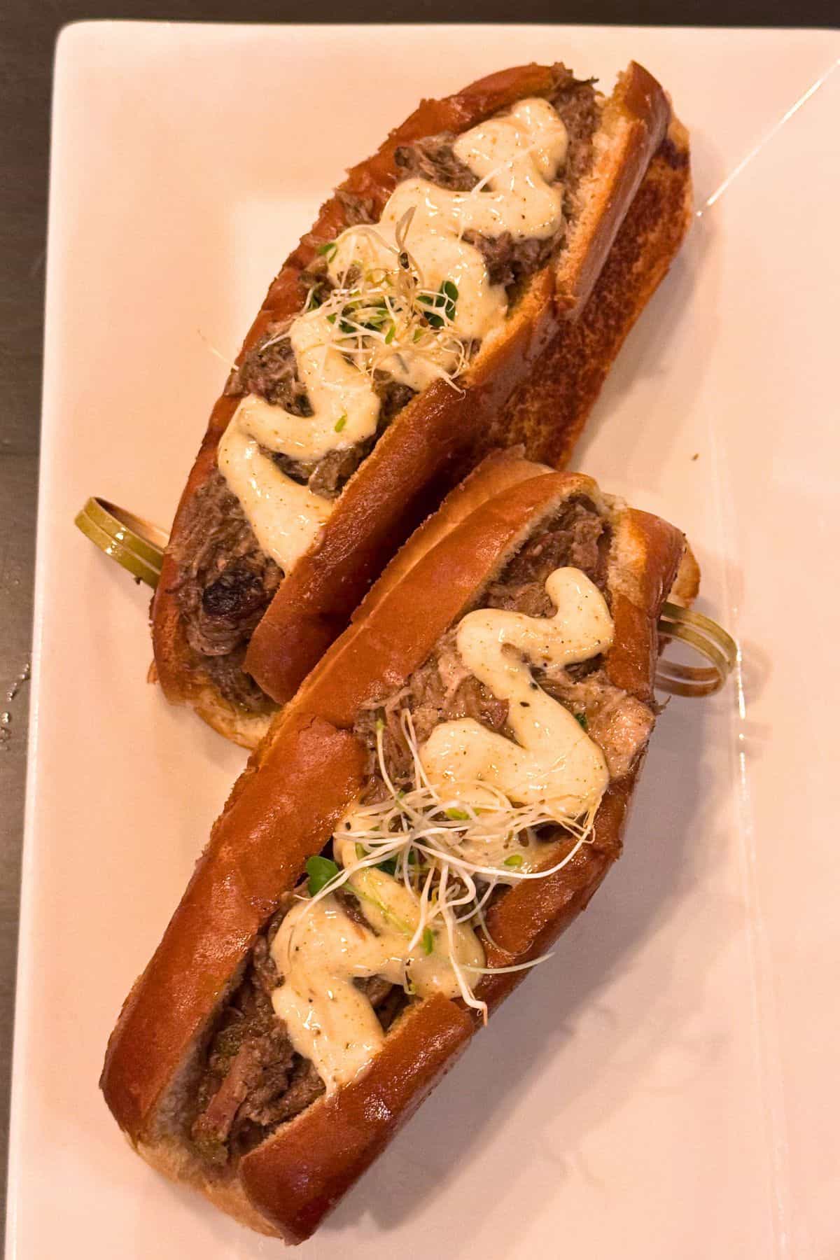 Two sandwiches on a white plate, each filled with sliced meat, creamy sauce, and topped with sprouts.