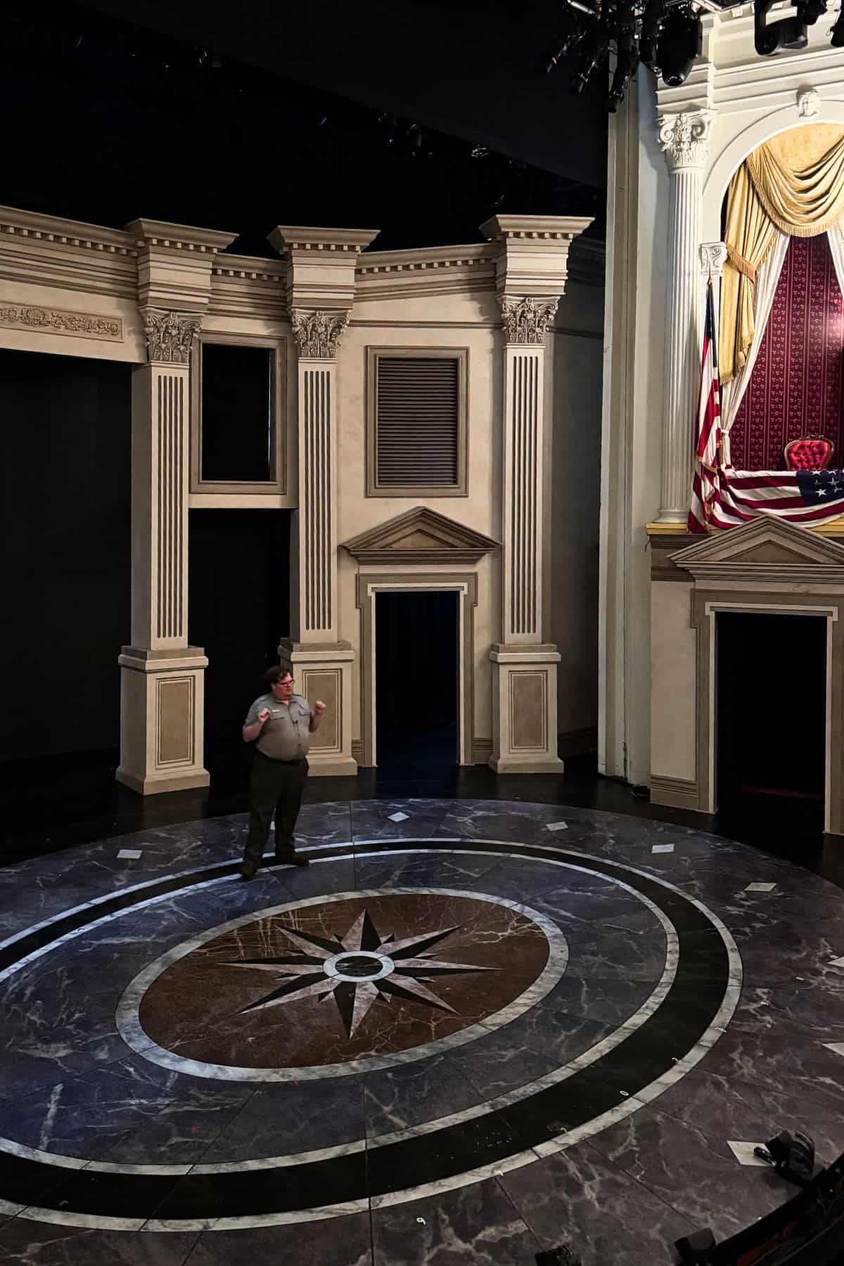 A person stands on stage near a circular floor design in front of a replica of historic architectural columns and a curtained balcony with American flags.