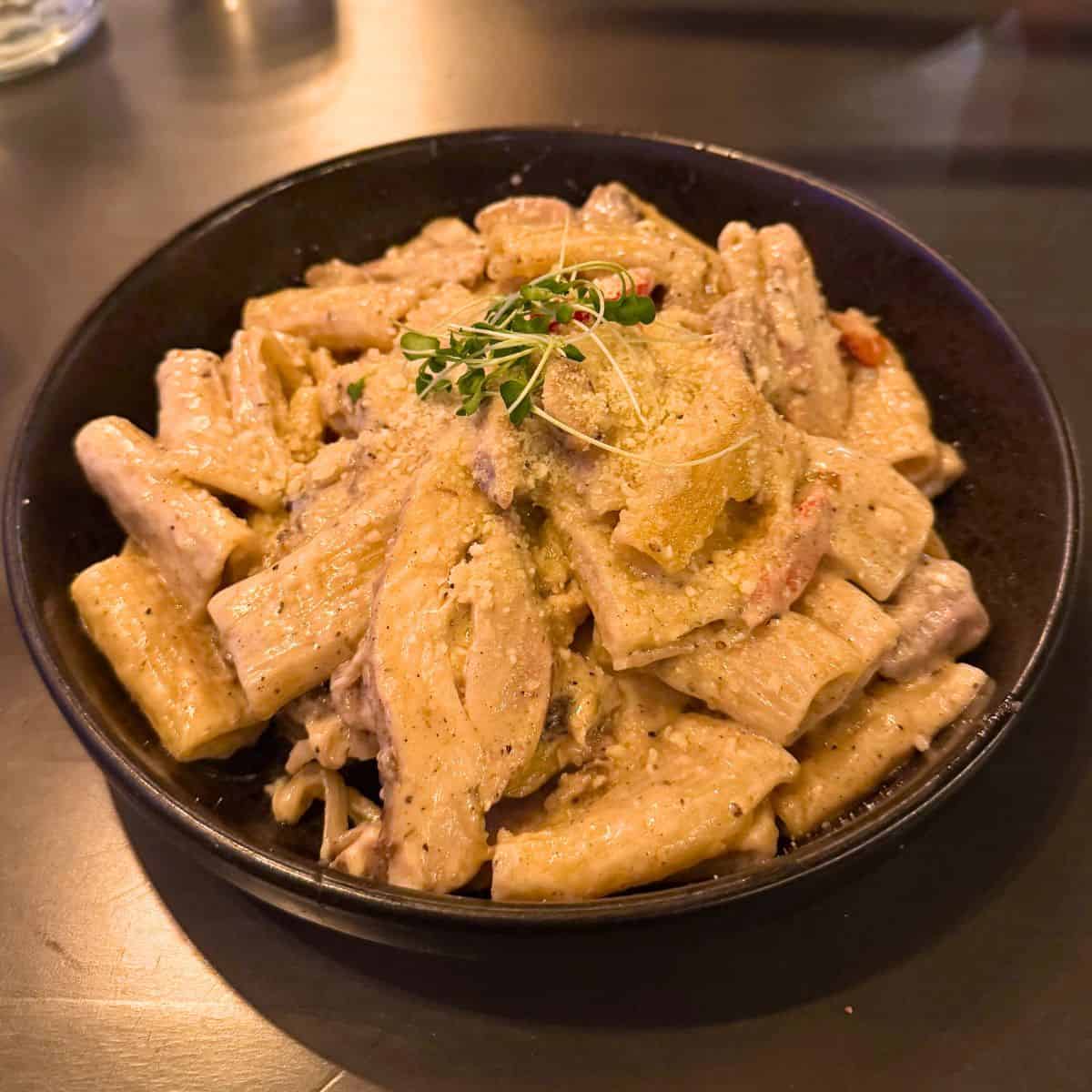 A bowl of rigatoni pasta with creamy sauce, chicken slices, herbs, and grated cheese, served on a dark plate.
