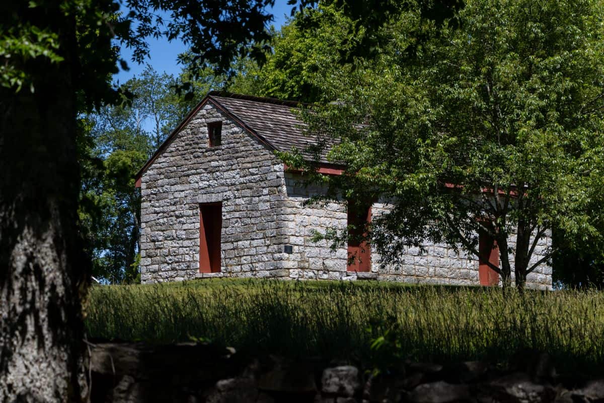 A small stone building with red doors and a shingled roof sits on a grassy hill, surrounded by green trees under a clear blue sky.