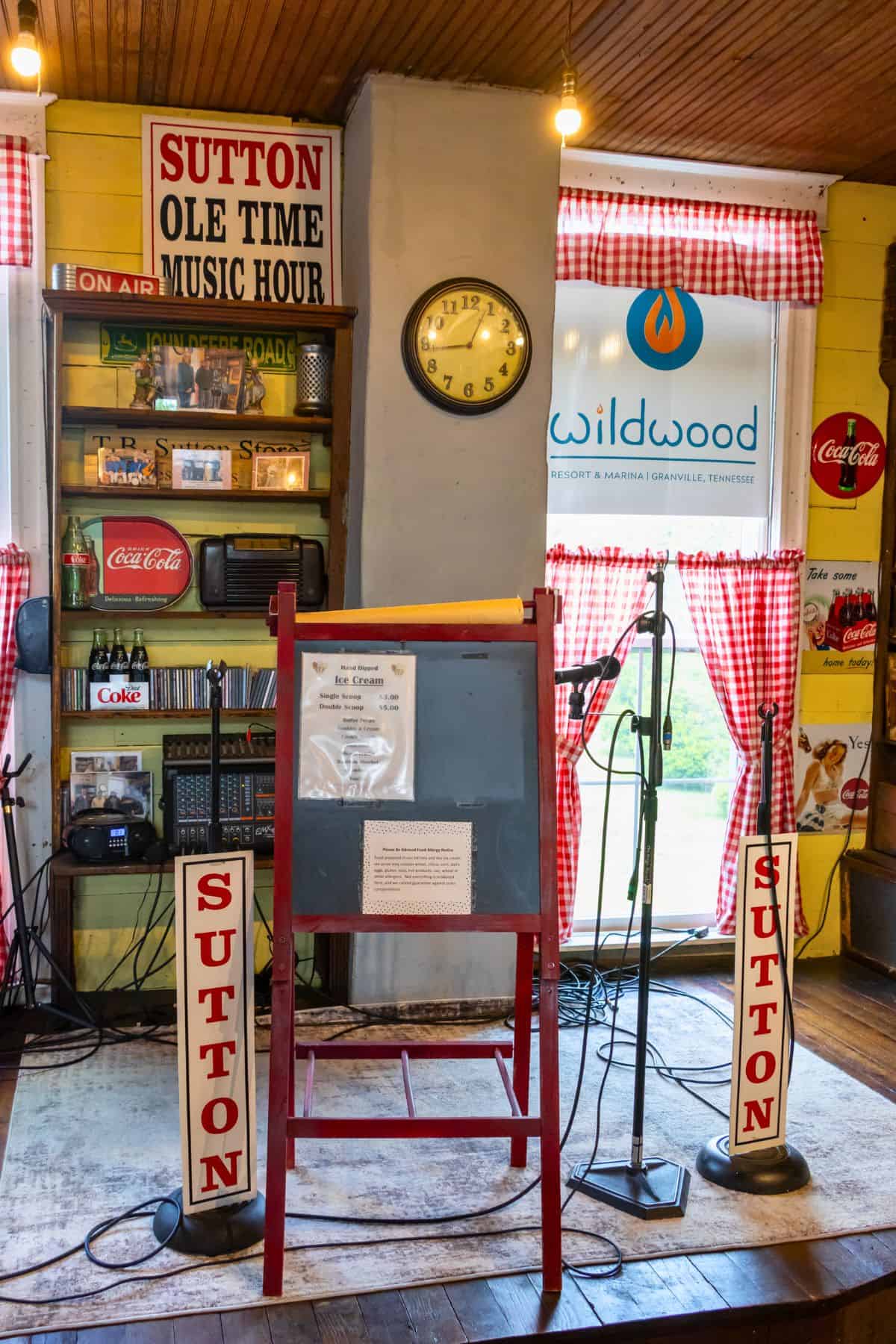 A small stage setup with microphones, speakers, vintage Coca-Cola signs, and "Sutton Ole Time Music Hour" signs, under a clock and a window with red checkered curtains.