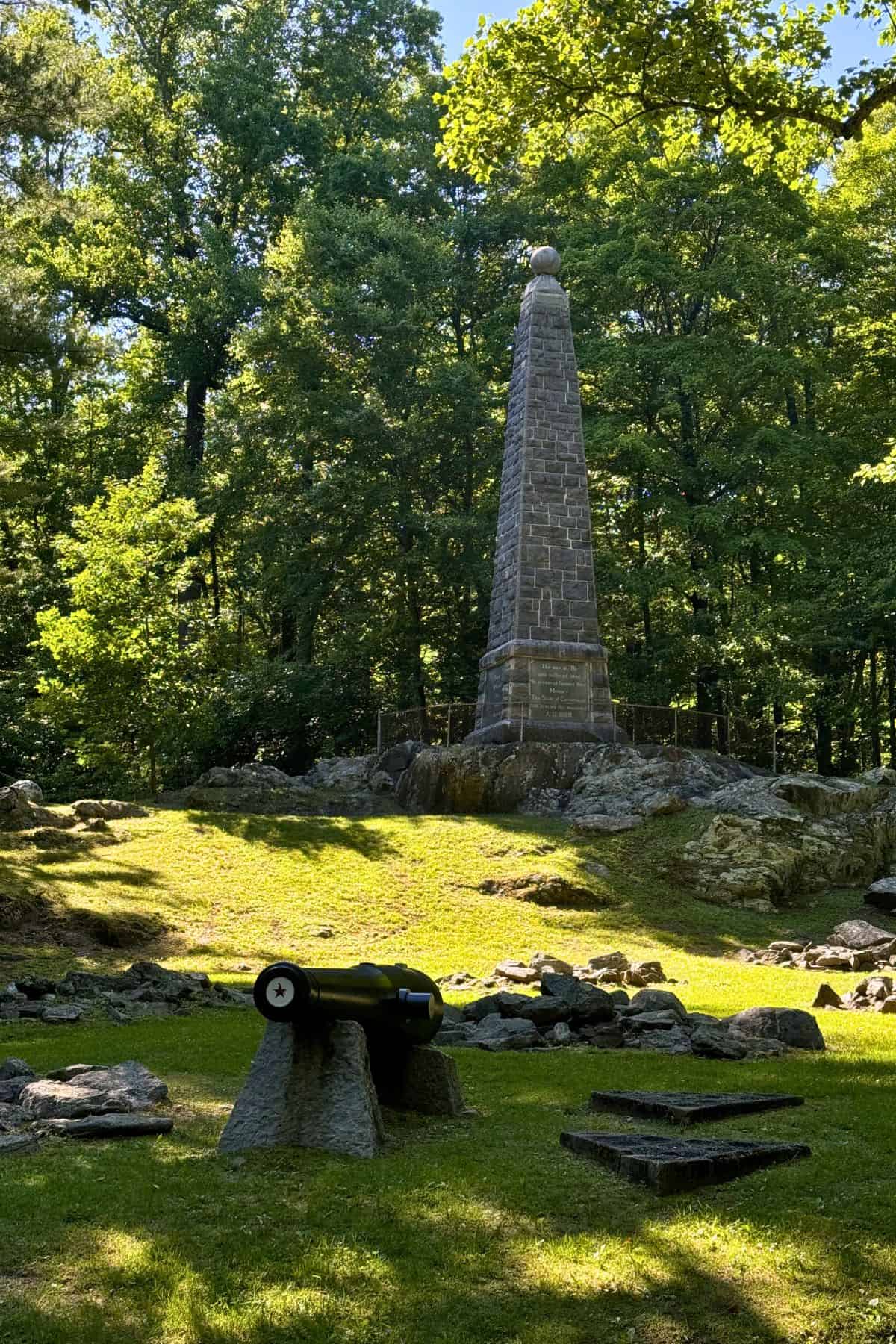 A stone obelisk monument stands in a grassy clearing with a historical cannon in the foreground, surrounded by trees and scattered rocks.