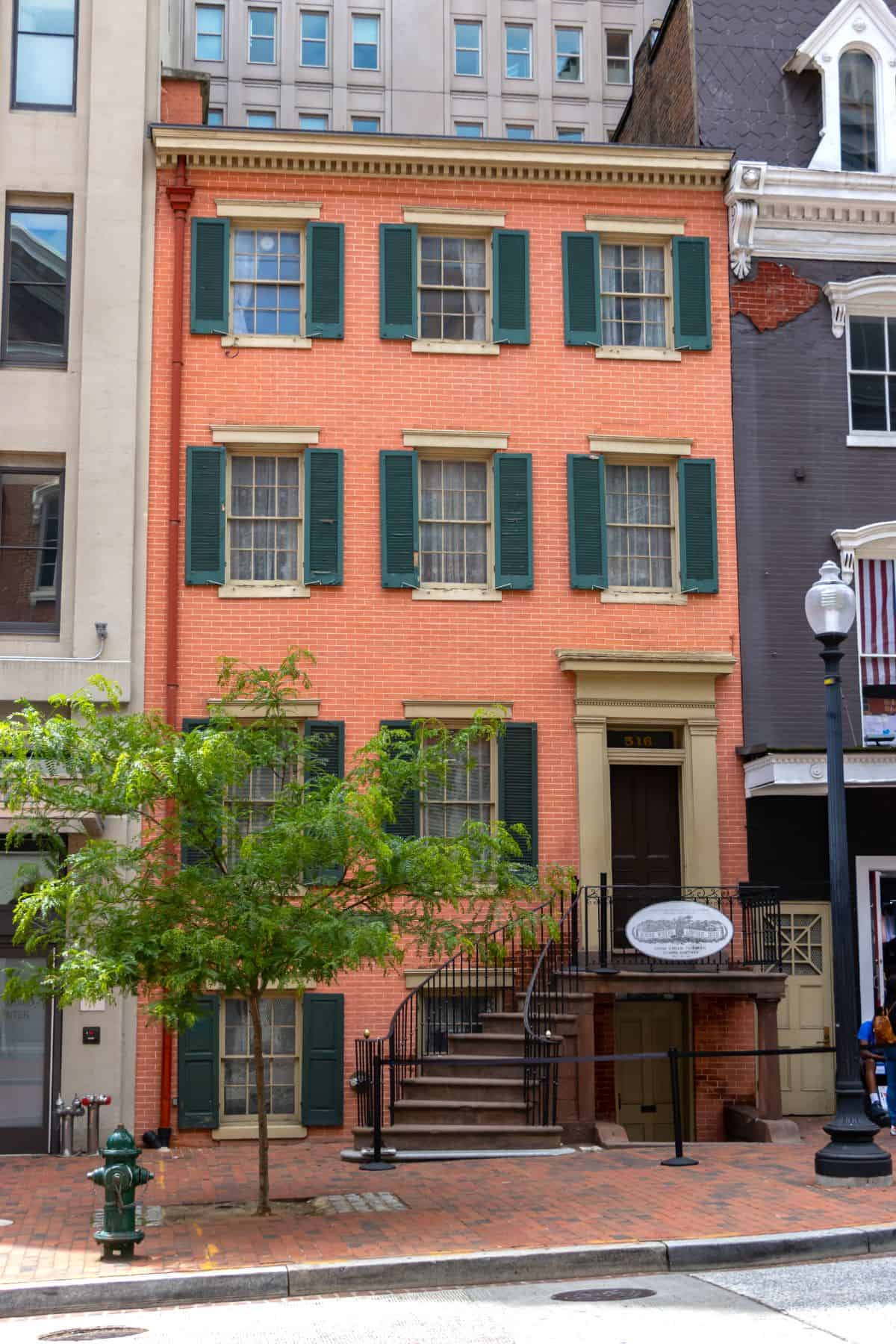 Three-story brick rowhouse with green shutters, arched doorway, iron railings, and a plaque near the entrance, located on a city street with a tree in front.