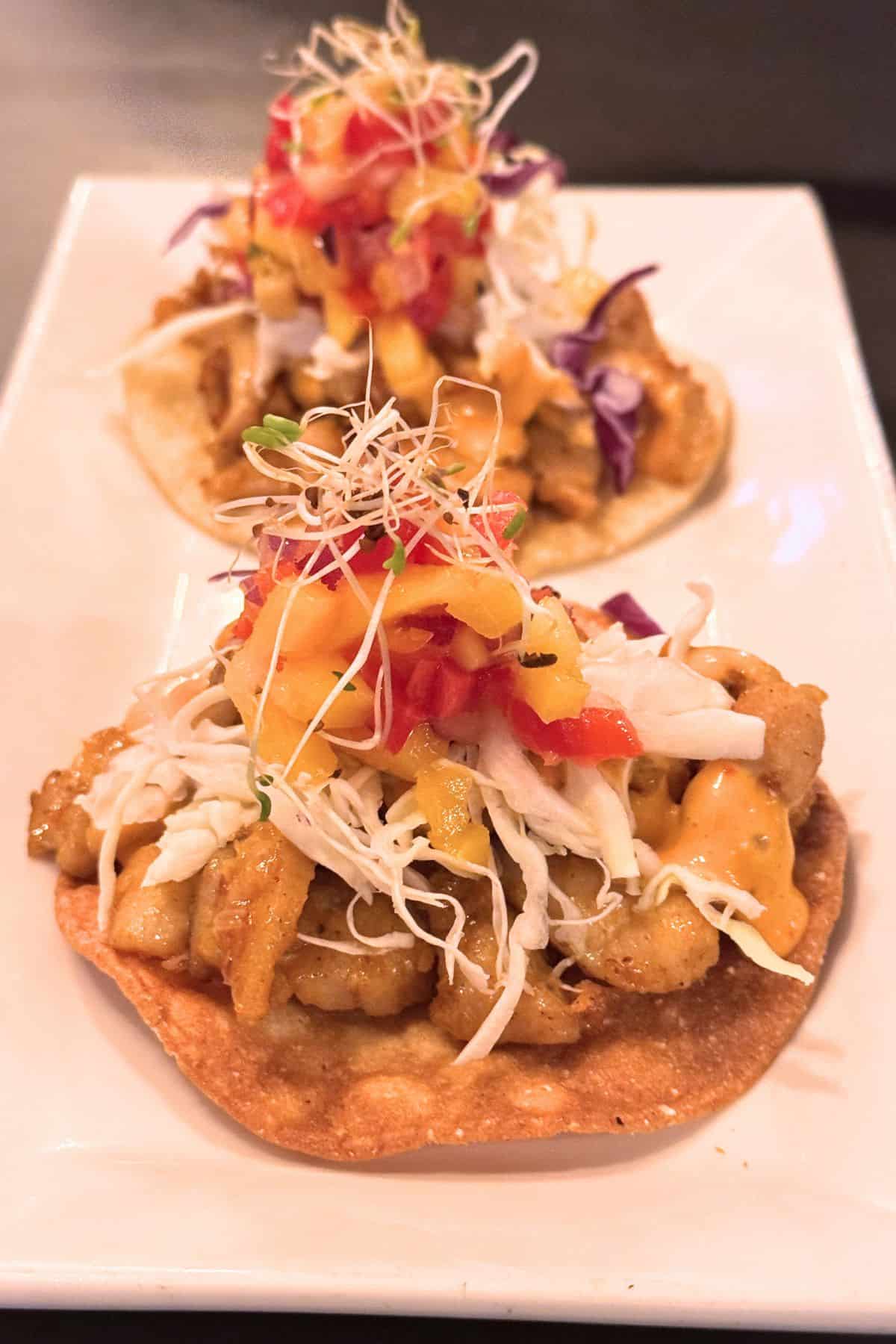 Two tostadas topped with sautéed vegetables, shredded cabbage, diced tomatoes, sprouts, and sauce are served on a rectangular white plate.