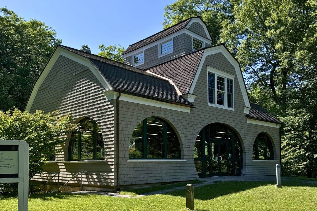 A gray shingle building with large arched windows, a steep roof, and a cupola, surrounded by green trees on a sunny day.