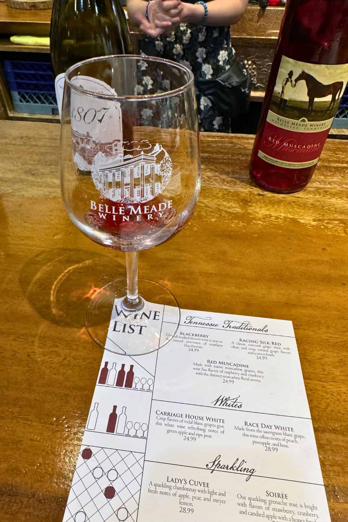 A wine tasting setup at Belle Meade Winery with a wine glass, two bottles of wine, and a wine list on a wooden counter.