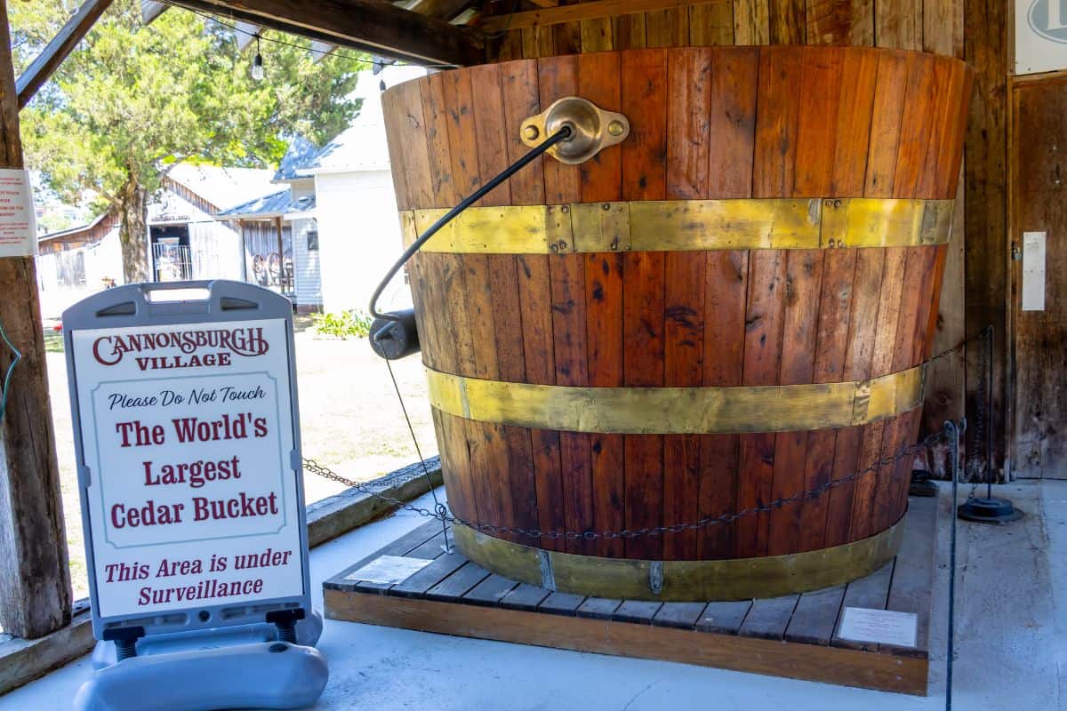 A large wooden bucket is displayed outdoors at Cannonsburgh Village, with a sign stating it is the world's largest cedar bucket and that the area is under surveillance.