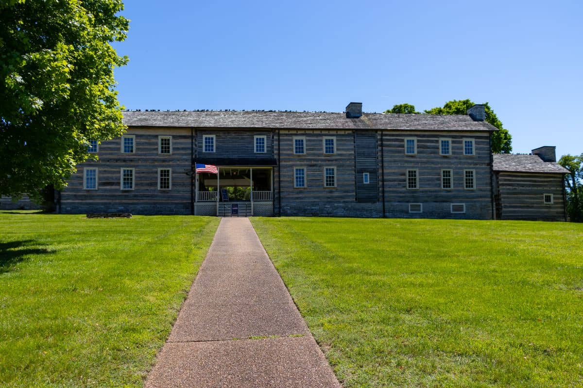 A wide, two-story log building with an American flag, set on a lawn with a paved walkway leading to its entrance under a clear blue sky.