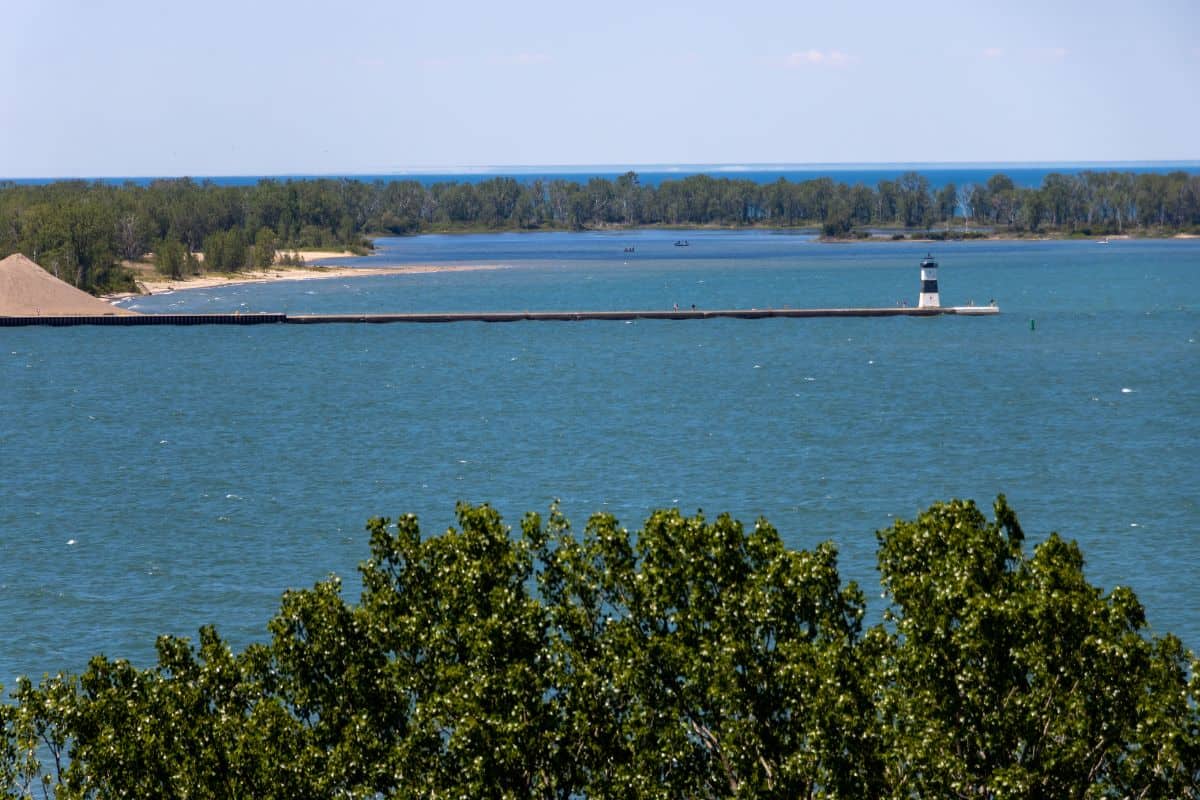 A body of water with a long breakwater, a small lighthouse, trees in the foreground, and forested land in the background under a clear sky.