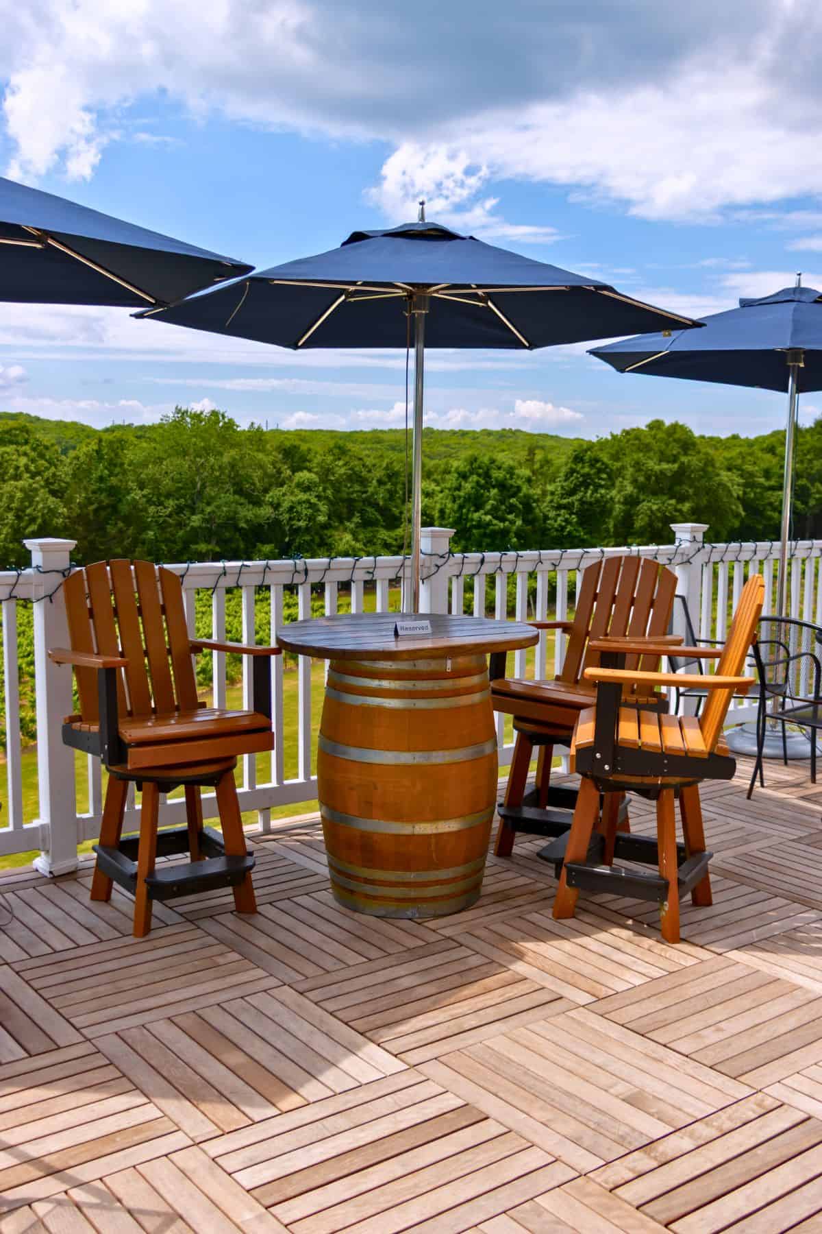 Two wooden chairs and a barrel table sit under a large outdoor umbrella on a wooden patio with a white railing and green trees in the background.