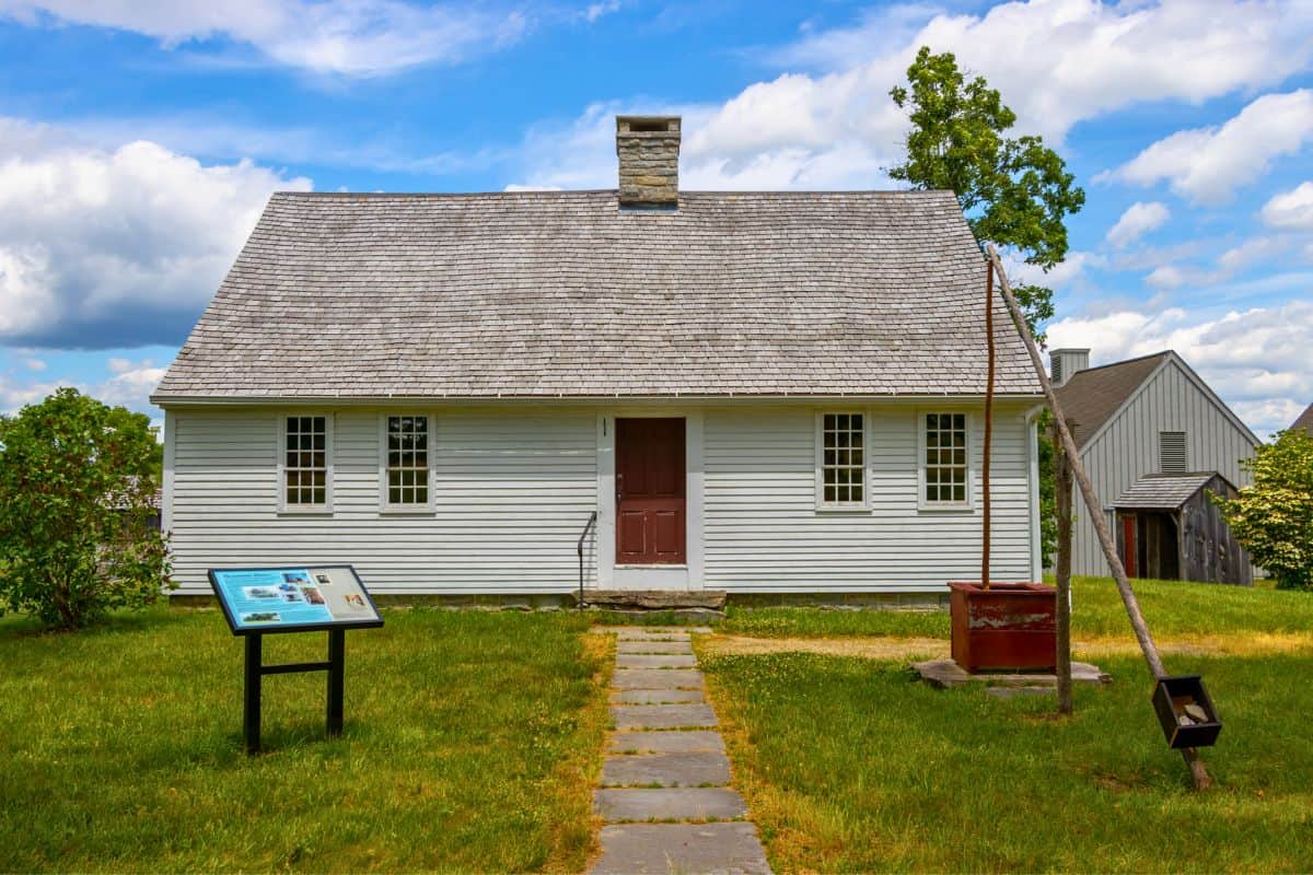 A historic white wooden house with a stone path leading to the door, an information sign, a well, and outbuildings in the background under a partly cloudy sky.