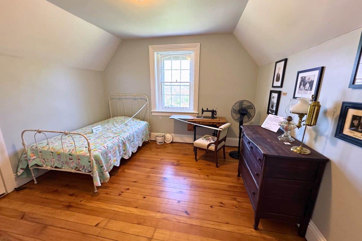A small bedroom with a single metal bed, sewing machine, wooden dresser, fan, lamp, framed pictures on the wall, and a window letting in natural light.