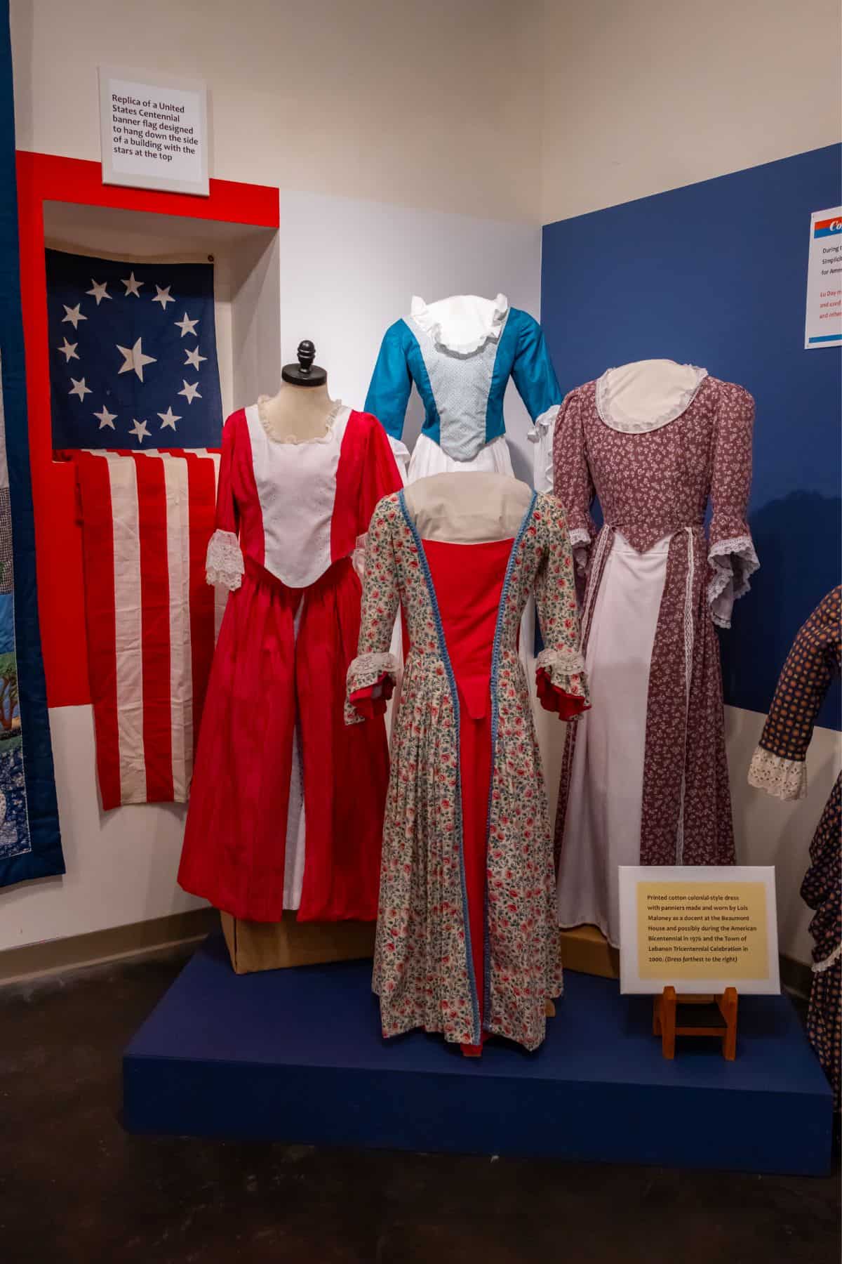 Exhibit display of three historical women’s dresses and an early American flag, with informational plaques on the wall and stand.