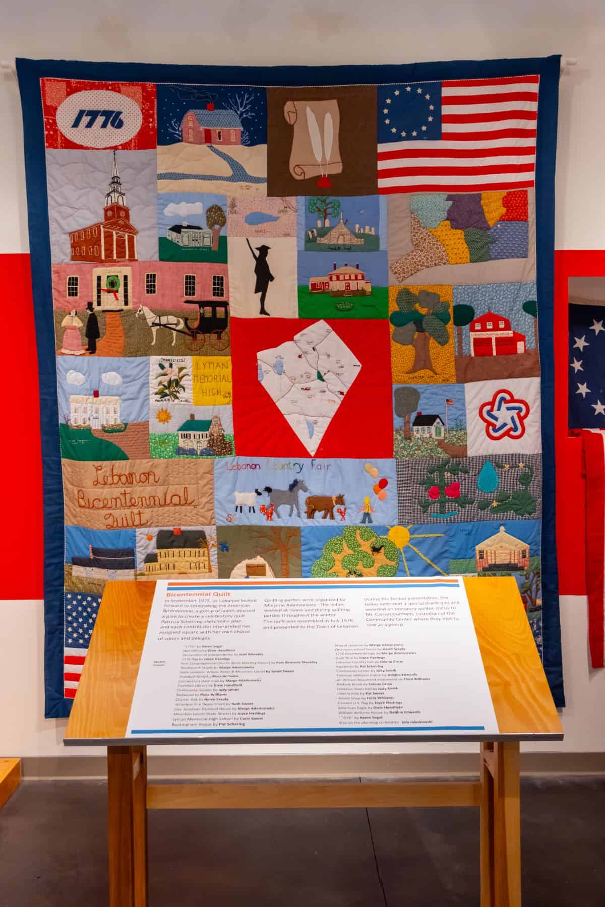 A colorful patchwork quilt featuring historic buildings, American flags, and symbolic images, displayed on a wall above an informational plaque on a wooden stand.
