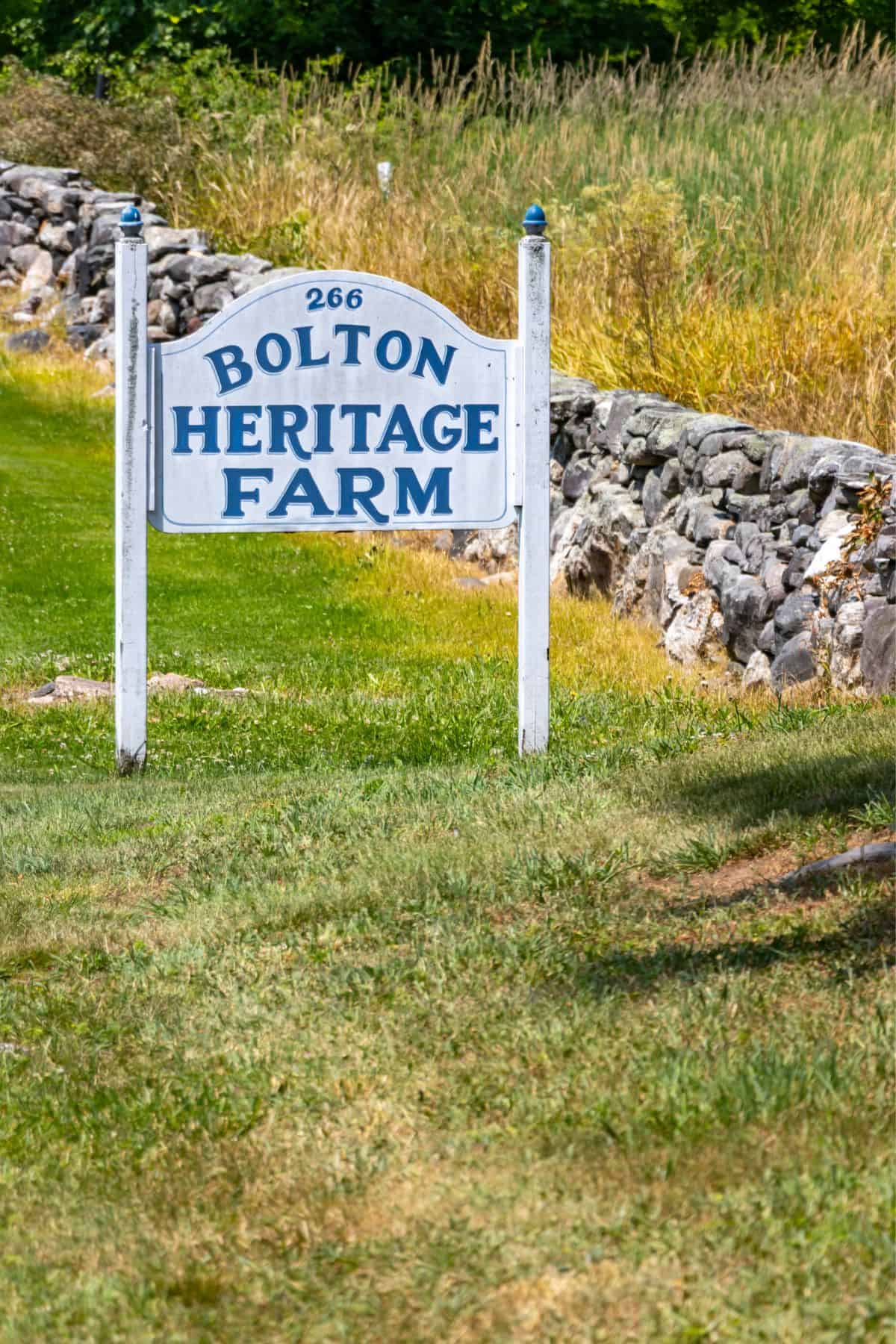 White sign reading “266 Bolton Heritage Farm” stands on a grassy area near a stone wall, with tall grass and trees in the background.