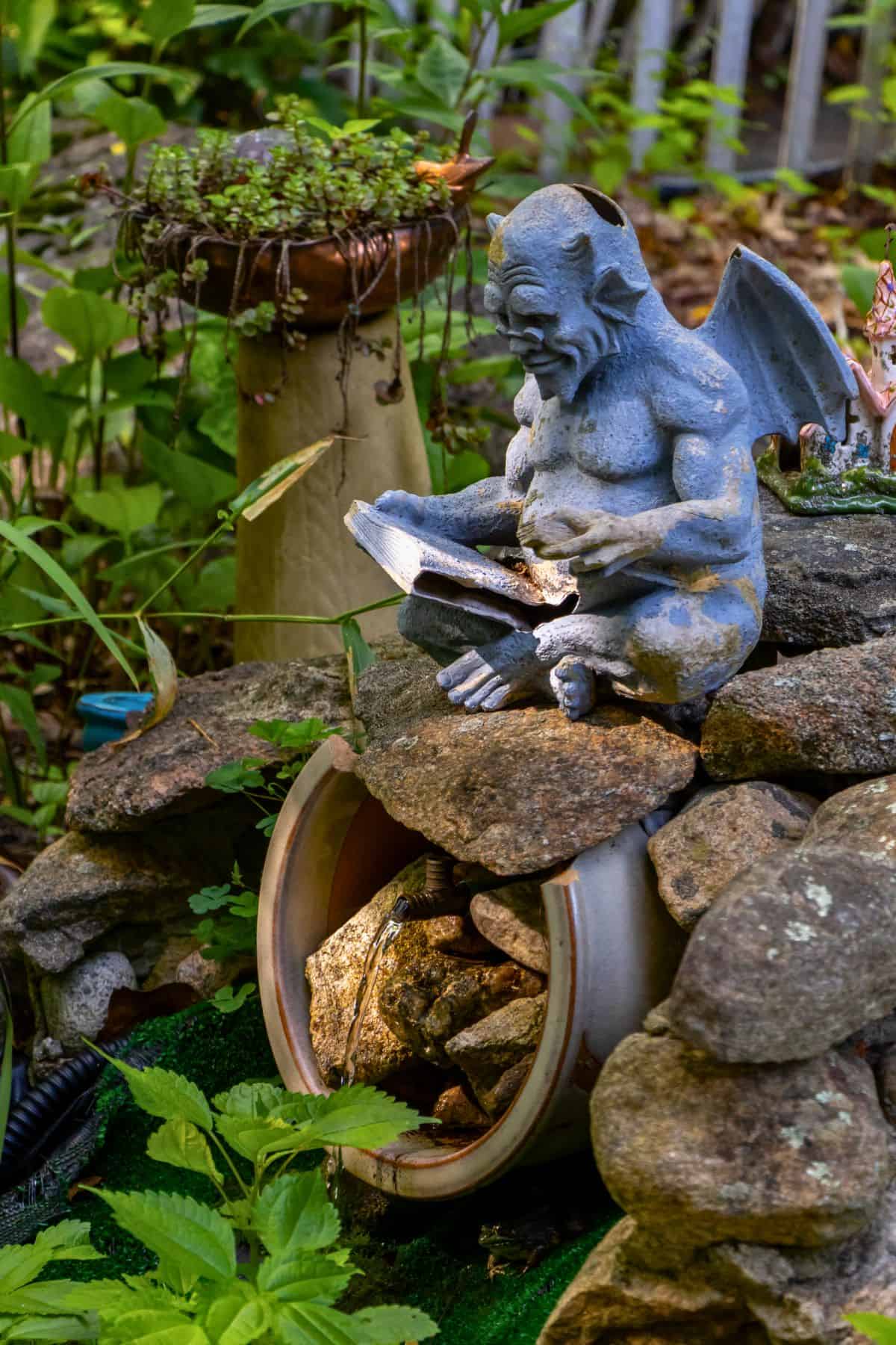 A stone gargoyle figurine with wings sits on rocks, reading a book, next to an overturned pot filled with stones in a garden setting.