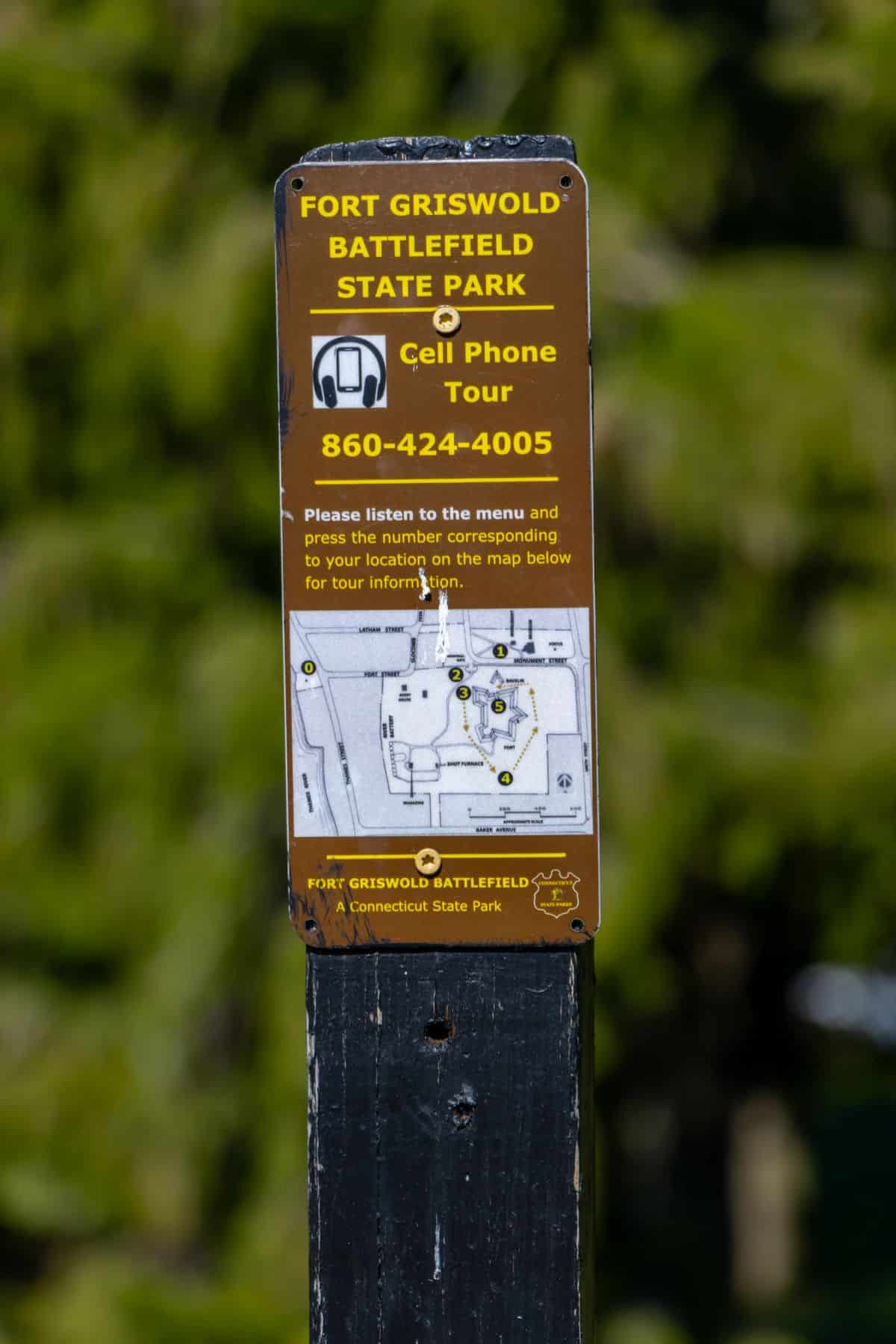 Brown sign for Fort Griswold Battlefield State Park showing a cell phone tour number, instructions, and a map of the park, mounted on a black post with trees in the background.