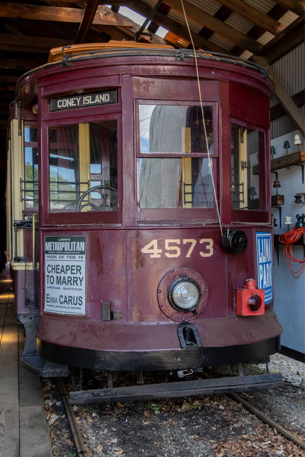 Vintage maroon streetcar numbered 4573 with "Coney Island" sign, parked indoors; features advertisements and old-fashioned design elements.