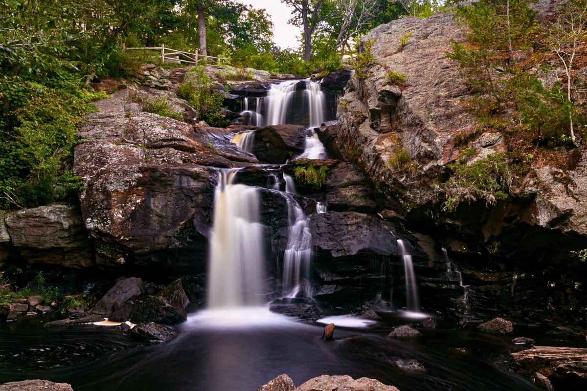 A multi-level waterfall flows over dark rocks surrounded by green trees, with a wooden fence visible at the top of the falls.