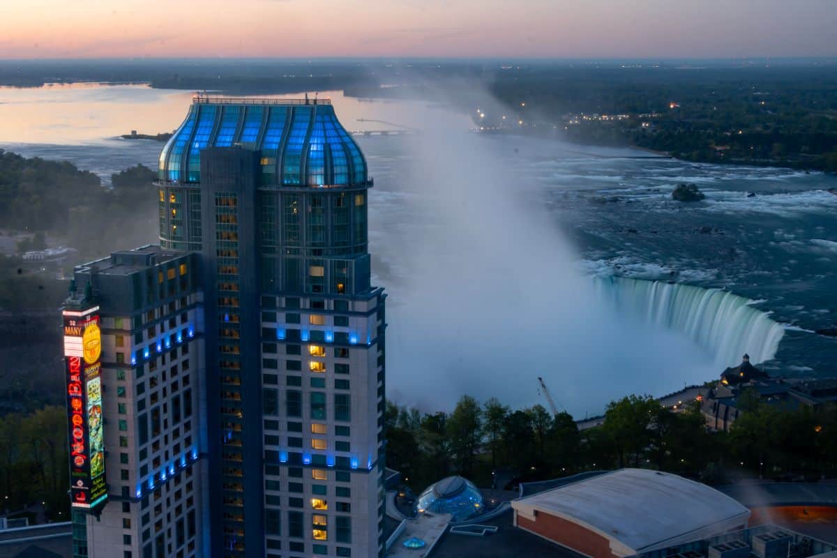 A tall hotel building stands near Niagara Falls at dusk, with mist rising from the waterfall and a river visible in the background.