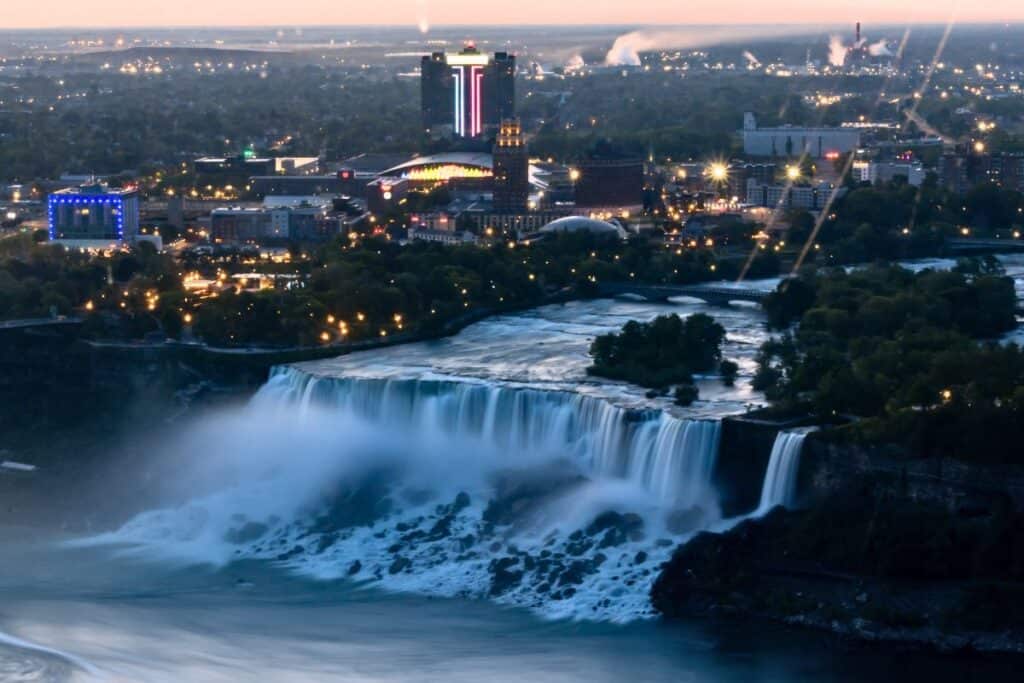 Aerial view of Niagara Falls with city buildings illuminated in the background at dusk.