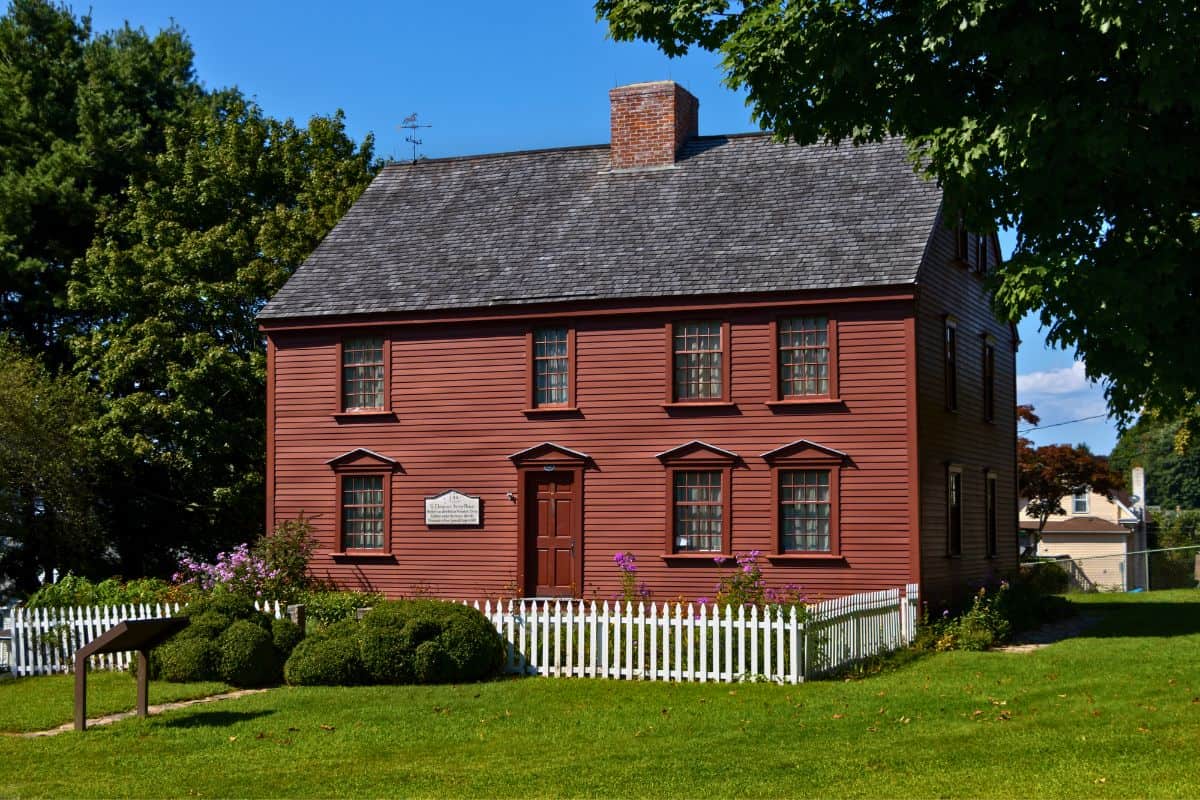 A two-story red wooden colonial house with a shingled roof, white picket fence, and neatly trimmed bushes on a sunny day.
