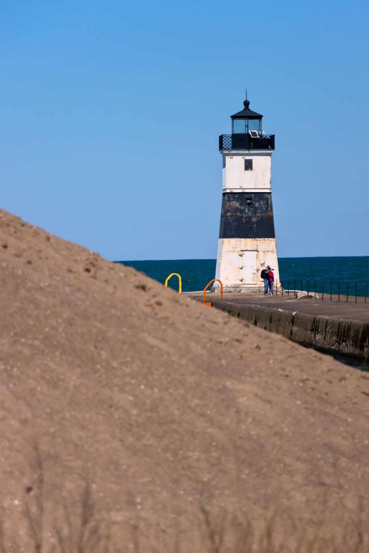 A black and white lighthouse stands on a pier by the water, with two people near its base and a sandy hill in the foreground under a clear blue sky.