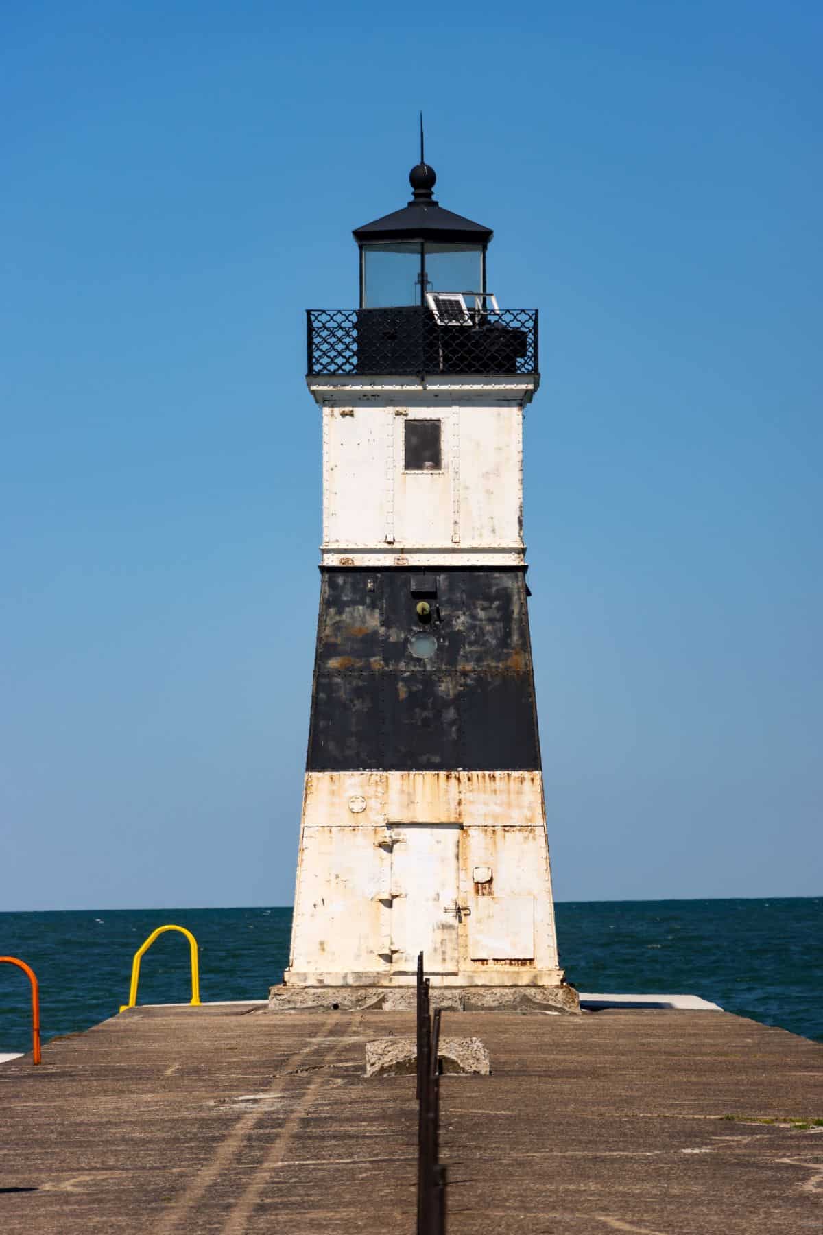 A weathered, black and white lighthouse stands at the end of a concrete pier, with the ocean and clear blue sky in the background.