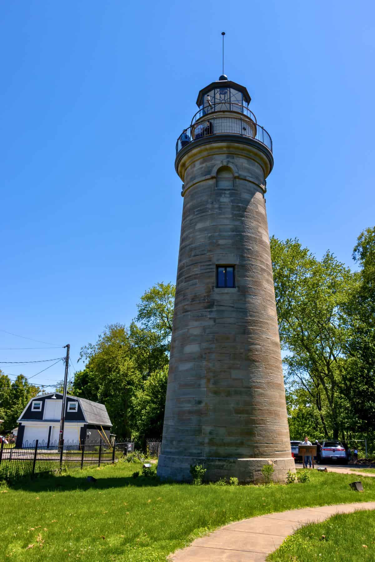 A tall, cylindrical stone lighthouse stands on a grassy lawn with trees and a small white building nearby under a clear blue sky.