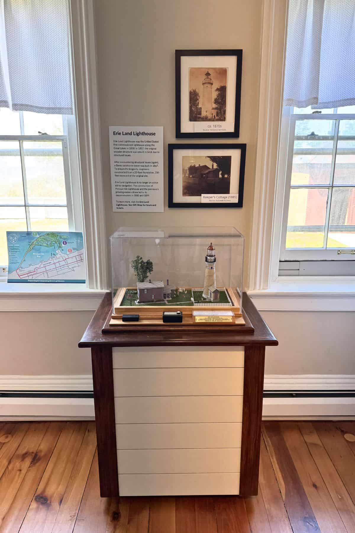 A display case with a model of the Erie Land Lighthouse sits on a cabinet, flanked by framed lighthouse photos and a map, in a room with wood floors and two windows.