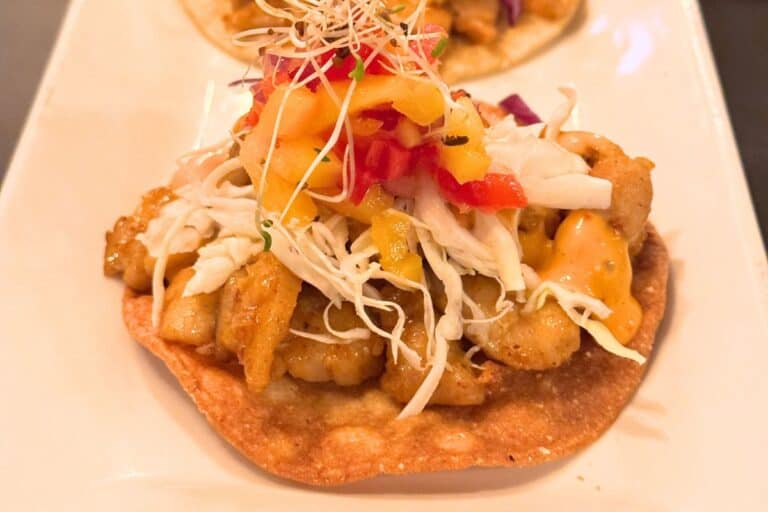 A crispy tostada topped with fried fish, shredded cabbage, diced tomatoes, mango salsa, and a drizzle of sauce, served on a white plate.