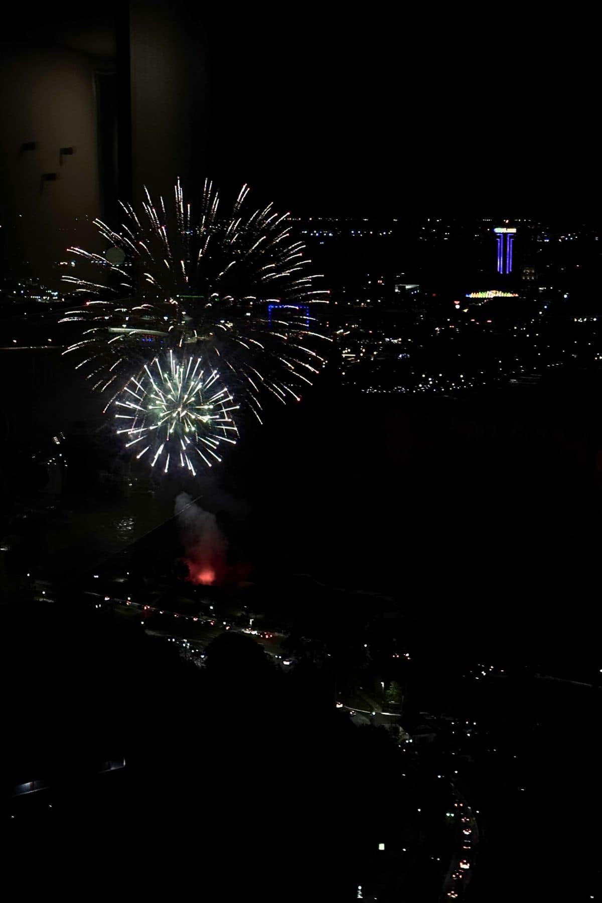Fireworks illuminate the night sky above a cityscape, with buildings and streetlights visible in the background.