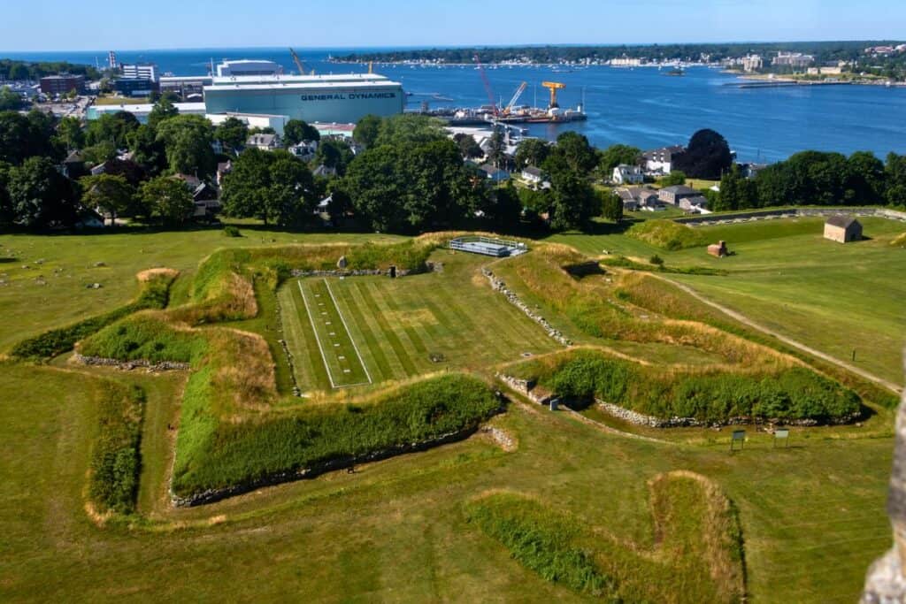 Aerial view of an old star-shaped fort with grassy earthworks near a river, surrounded by trees, with an industrial area and water in the background.