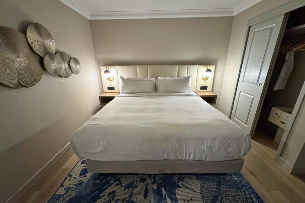 A neatly made bed with white linens, two bedside tables with lamps, decorative wall art, and an open closet in a neutral-toned bedroom.