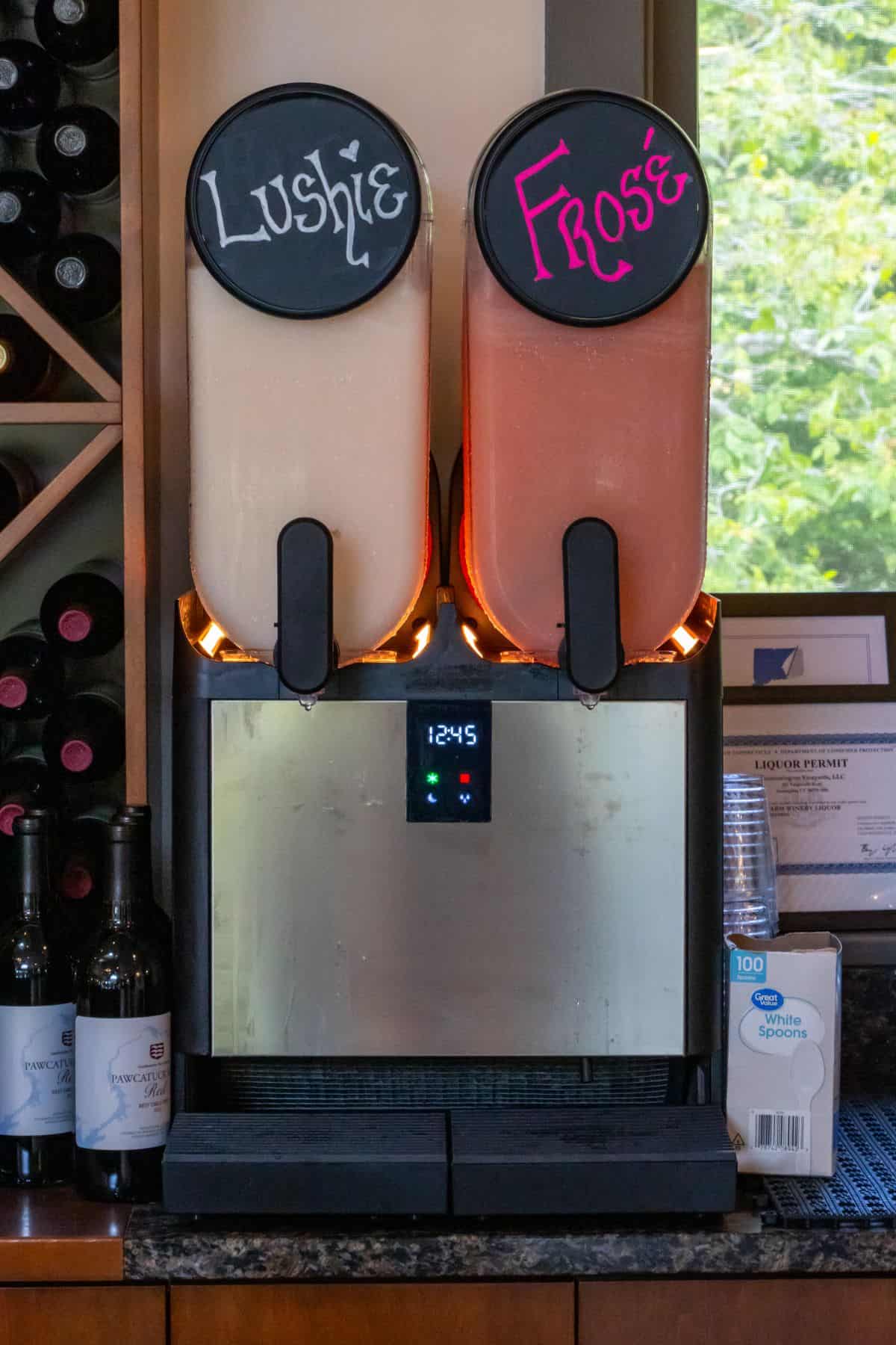 A beverage dispenser with two labeled sections, "Lushie" and "Frosé," is on a counter with wine bottles, cups, and plastic spoons nearby.