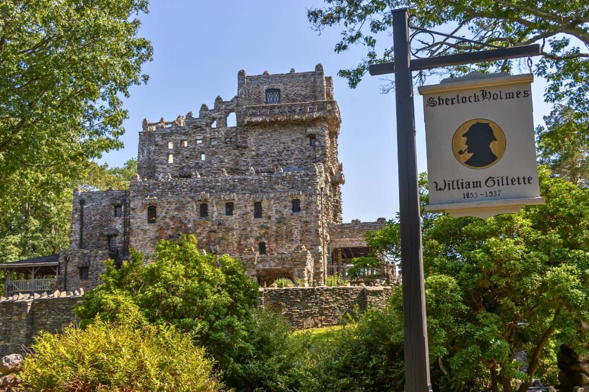 A stone castle partially obscured by trees, with a sign in the foreground that reads "Sherlock Holmes, William Gillette, 1853-1937.