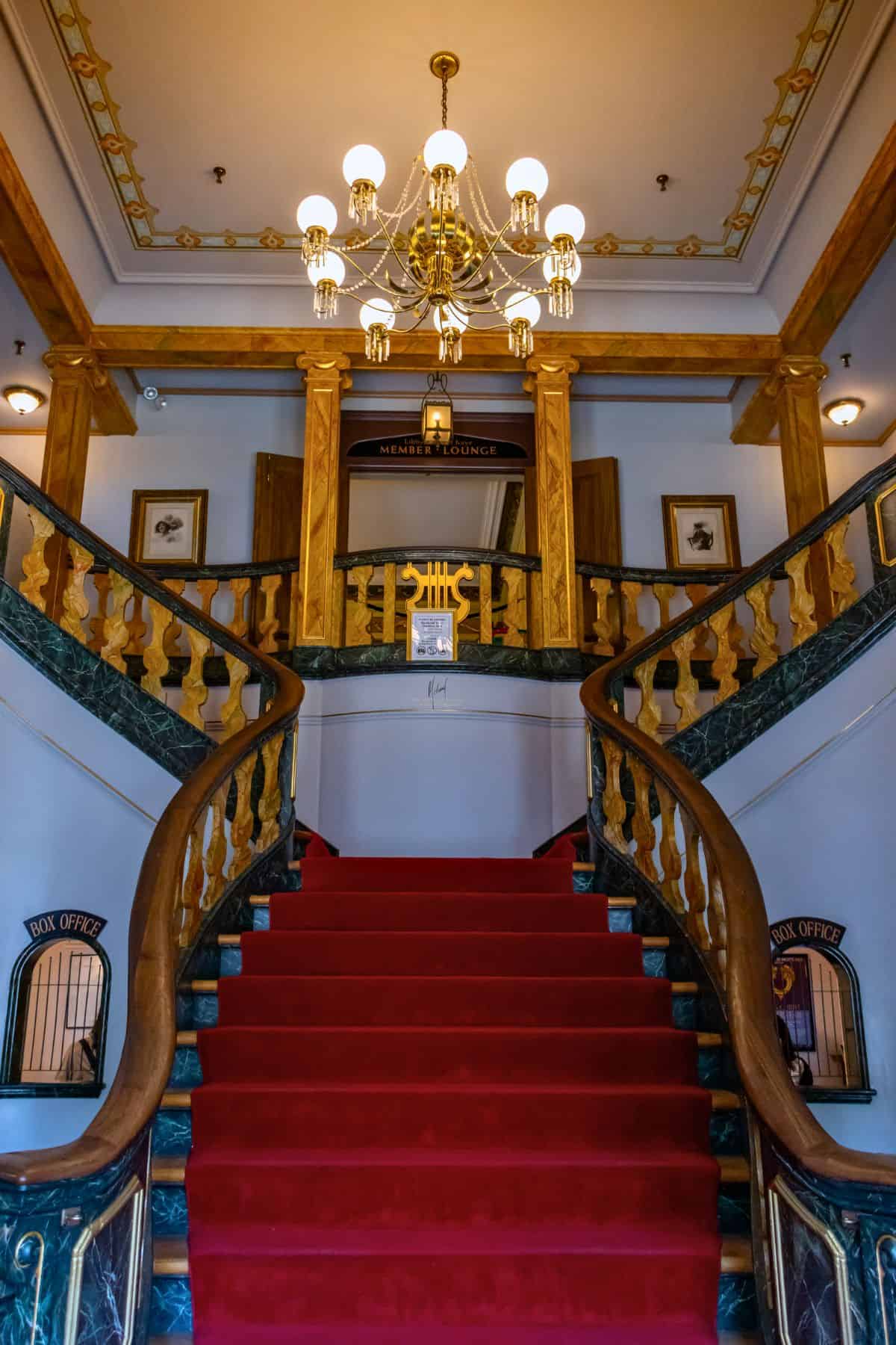 A grand staircase with a red carpet leads up to a wooden balcony, flanked by marble railings and lit by a chandelier in an ornate interior.