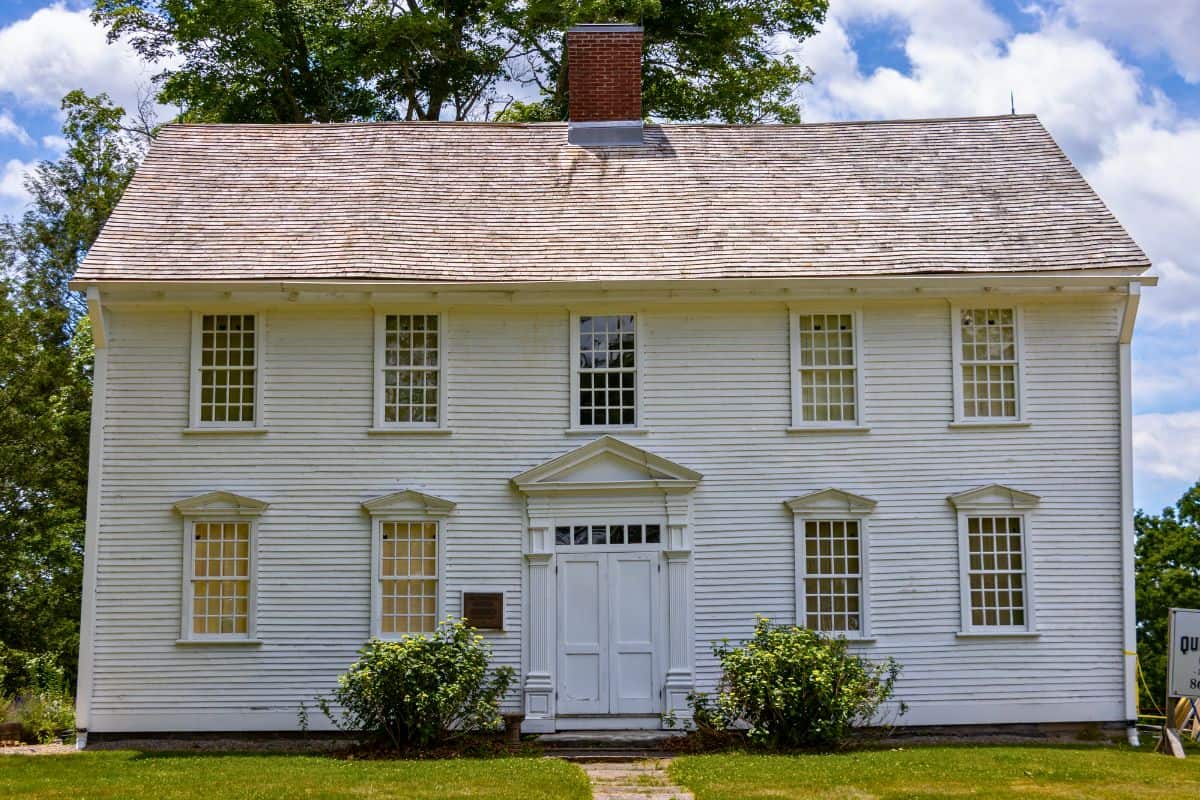 A two-story, white, wooden colonial-style house with many windows and a central chimney, surrounded by green shrubs and grass.