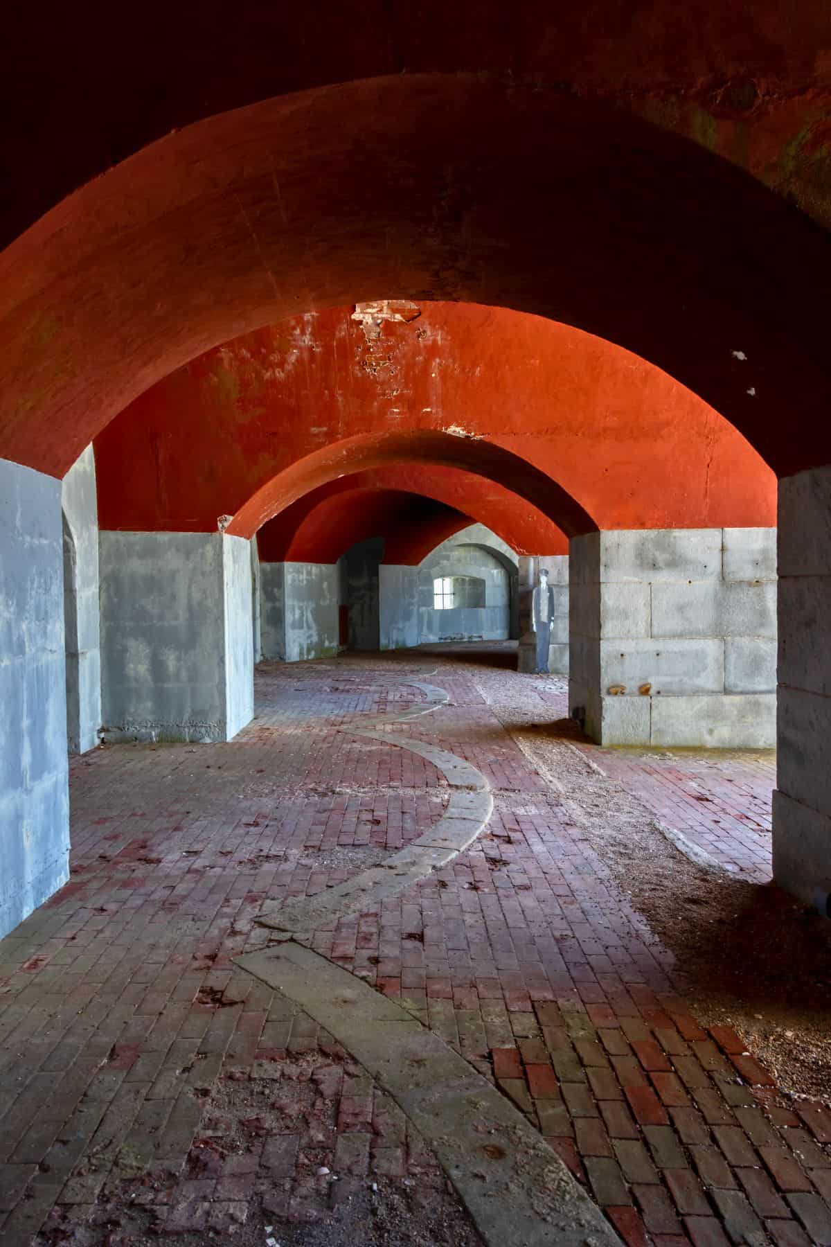 Interior view of an arched brick corridor with red vaulted ceilings, stone lower walls, and a winding pattern on the floor leading to windows in the distance.