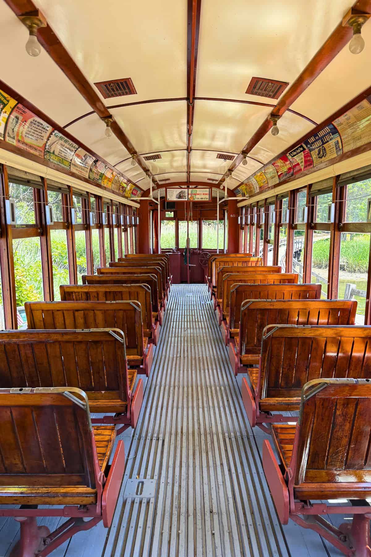Interior of an old-fashioned tram with wooden seats and vintage advertisements above the windows, viewed from the back toward the front doors.