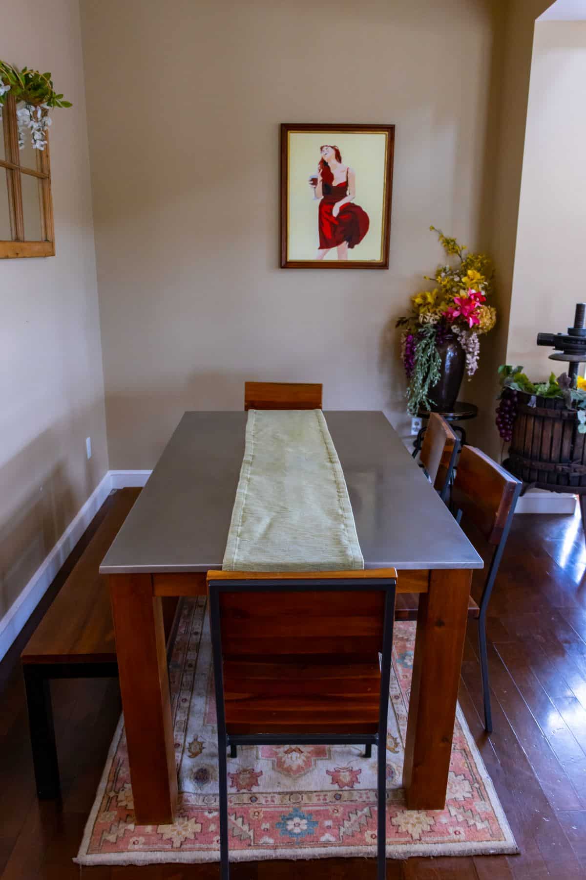 A dining area with a wooden table and four chairs, a runner on the table, a wall painting, a floral arrangement, and a decorative wine barrel in the corner.