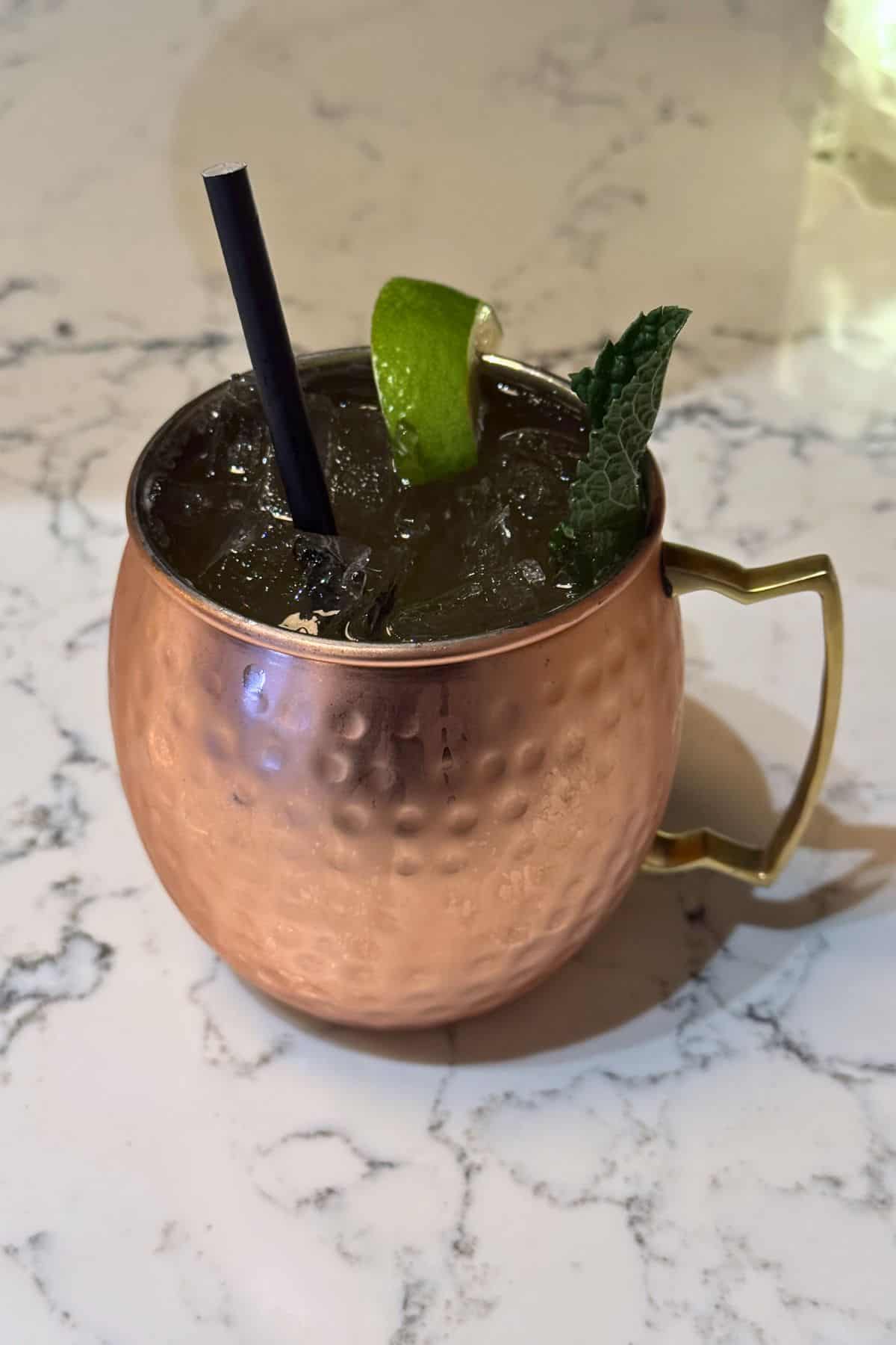 A copper mug filled with ice, garnished with a lime wedge and mint sprig, and served with a black straw on a white marble surface.