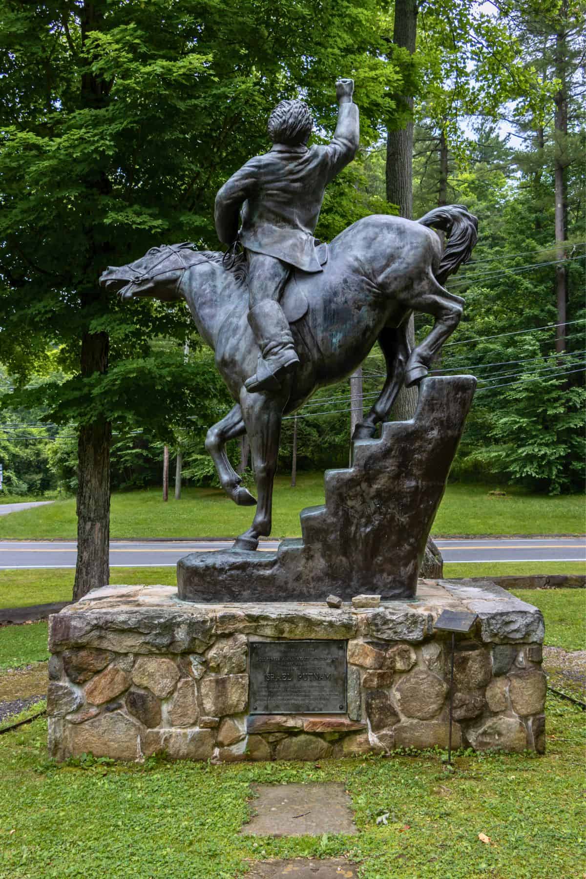 Bronze statue of a man on horseback ascending stone steps, mounted on a stone base, surrounded by trees and grass.