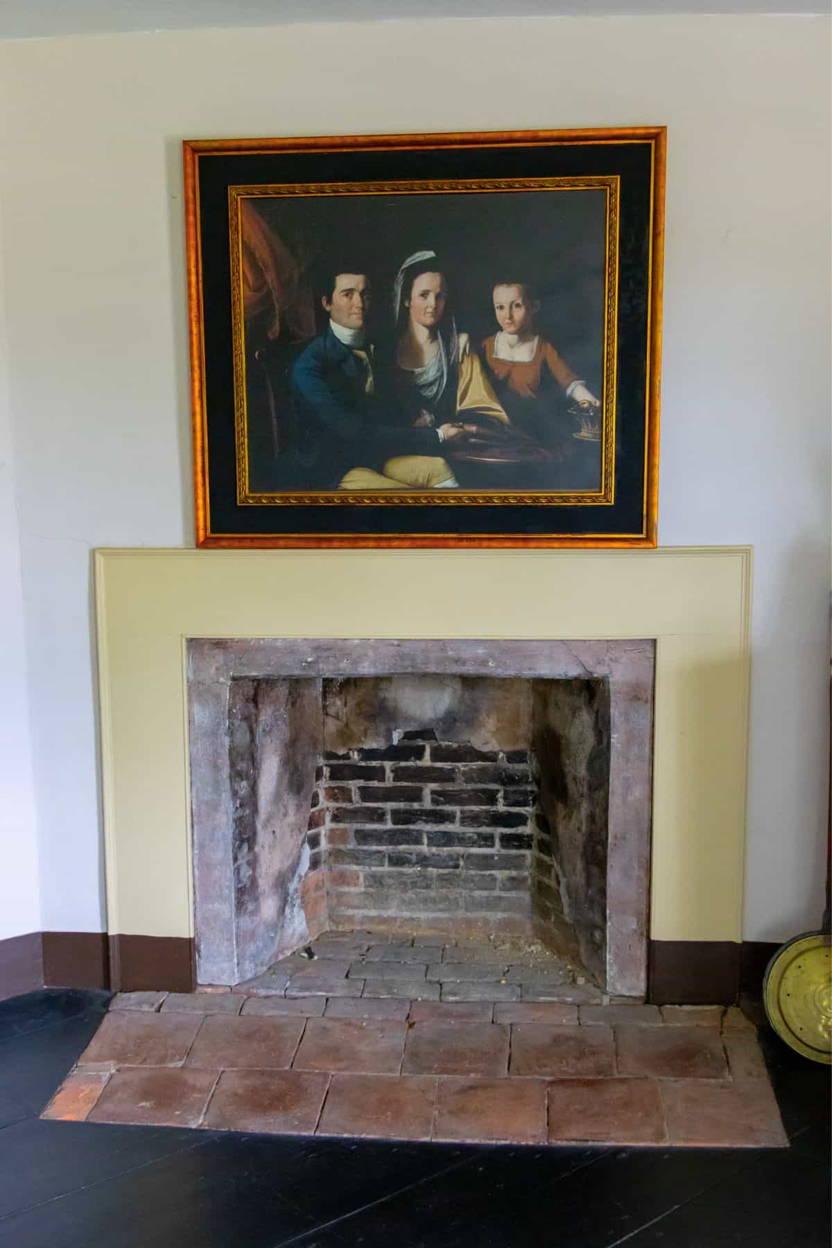 An empty brick fireplace is set into a wall with a large framed painting of three people hanging above it.