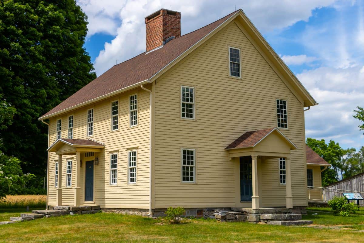 A two-story yellow wooden house with white trim, multiple windows, a brick chimney, and two covered entrances, surrounded by grass and trees under a partly cloudy sky.